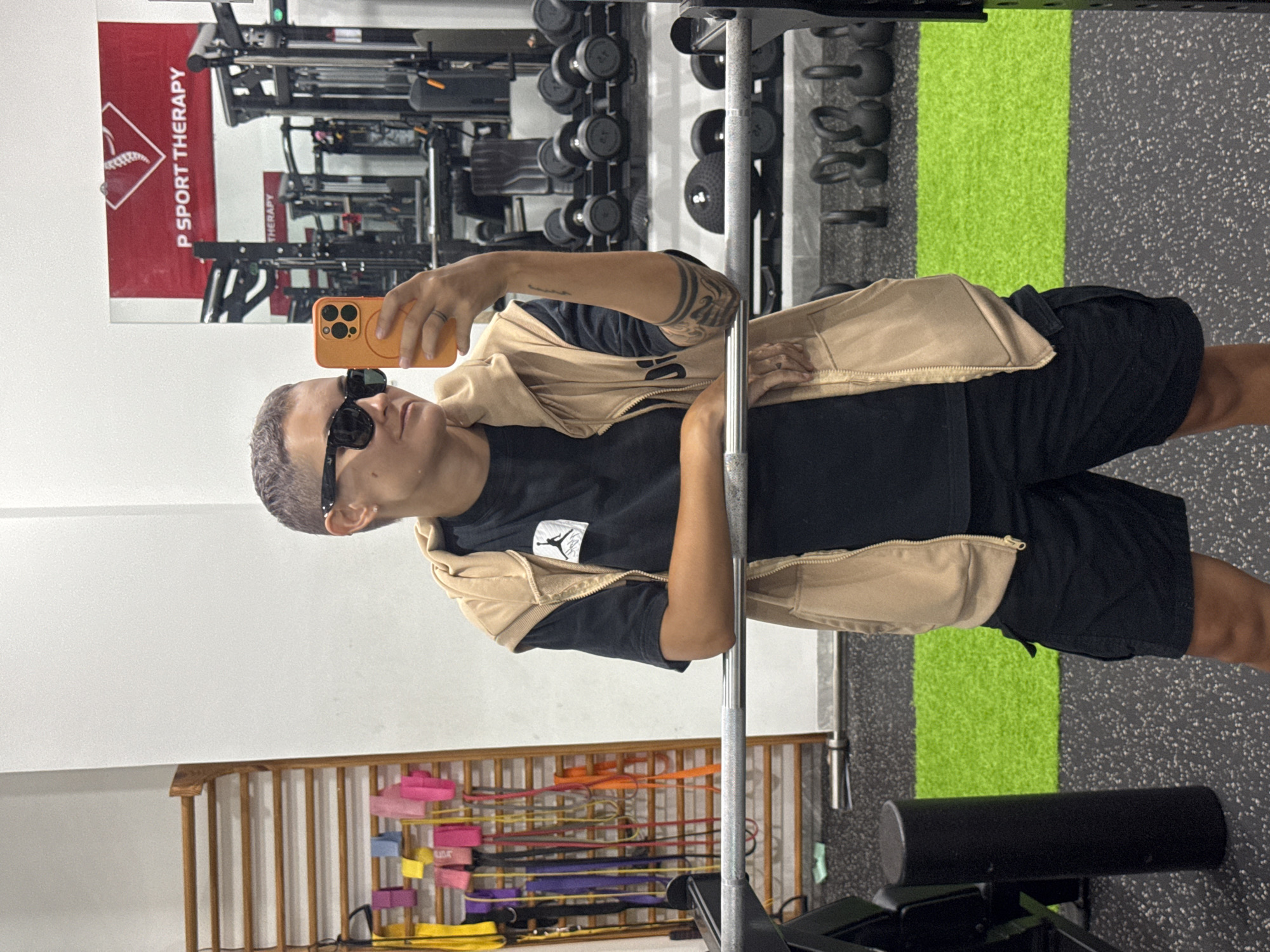 A person with short hair, wearing sunglasses, a tan vest over a black t-shirt and black shorts, is taking a selfie in a gym. The person is holding an orange smartphone in their right hand. The background shows gym equipment, including weights, a rack, and a wooden structure with resistance bands. A sign in the reflection of a mirror reads "P SPORT THERAPY". The floor is a speckled grey rubber mat with a bright green strip running through it. The scene appears to be indoors in a commercial gym. There is no information to place this image in the city of Nam Nha Trang Ward, Vietnam.