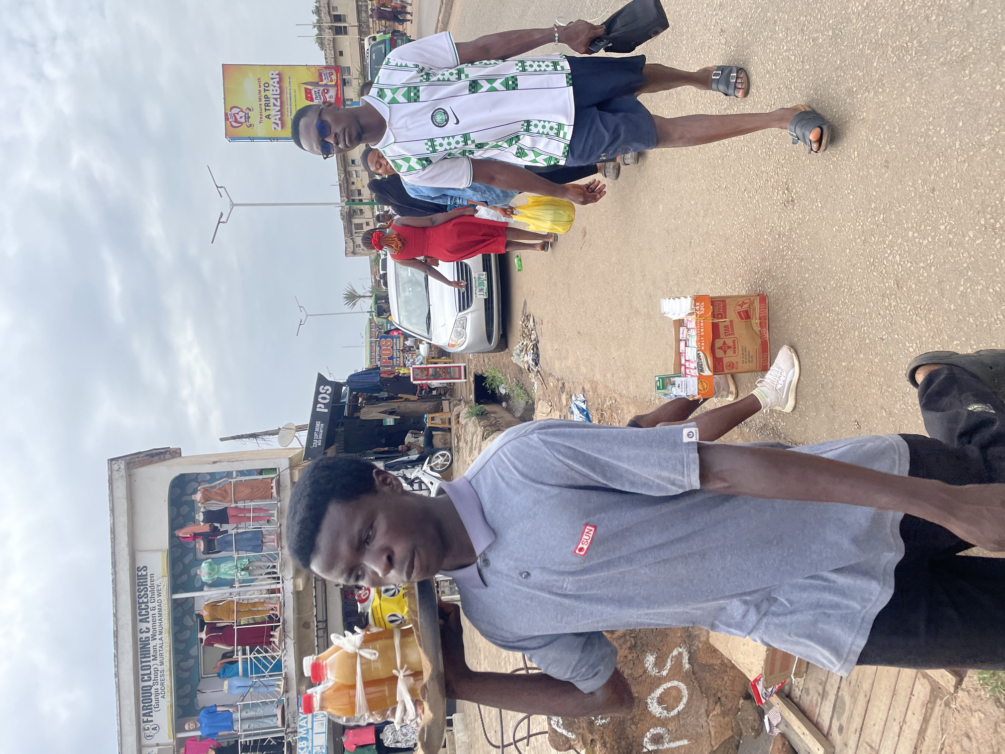The image depicts a street scene in Jos, Nigeria. A man in the foreground is carrying a tray of packaged orange juice, standing near a building labeled "FAROUQ CLOTHING & ACCESSORIES." The building also displays mannequins.
In the background, another man wearing a white and green soccer jersey is walking away from the camera, carrying a black bag. There are other people in the background, including a woman in a red dress, and a silver car with the license plate "JJN-382PU." Street signs, including one advertising a trip to Zanzibar, are visible. There are also beverage crates and POS signage nearby.