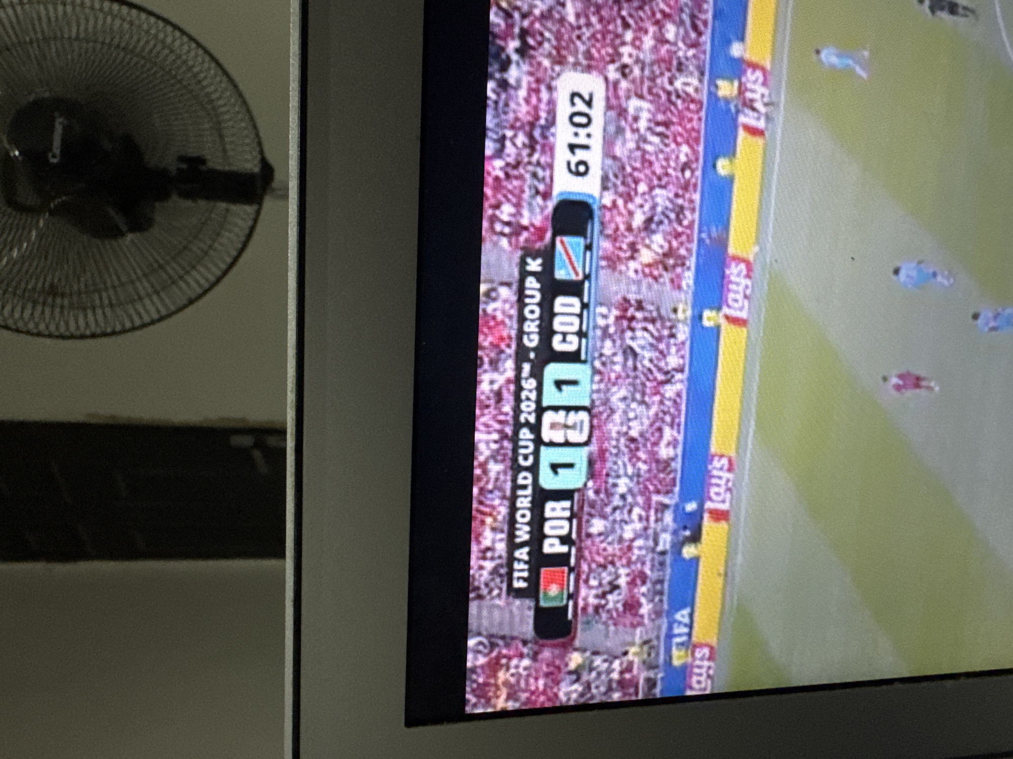 The image captures a television screen displaying a FIFA World Cup 2026 Group K match between Portugal (POR) and DR Congo (COD). The score is shown as 1-1, with the game clock at 61:02. The stadium's seating is filled with spectators in predominantly red attire, suggesting a large crowd. In the foreground of the screen, a green soccer field is visible with players in action. Advertising boards with "Lay's" branding are placed along the edge of the field. The television screen is positioned in front of a plain wall, with a black oscillating fan partially visible in the upper right corner of the frame, suggesting an indoor setting. The lighting is dim, with the television screen being the primary light source. The backdrop indicates a domestic viewing environment. The image does not contain information about Kufena, Nigeria, nor any identifiable landmarks or features that would place the scene in that specific location.