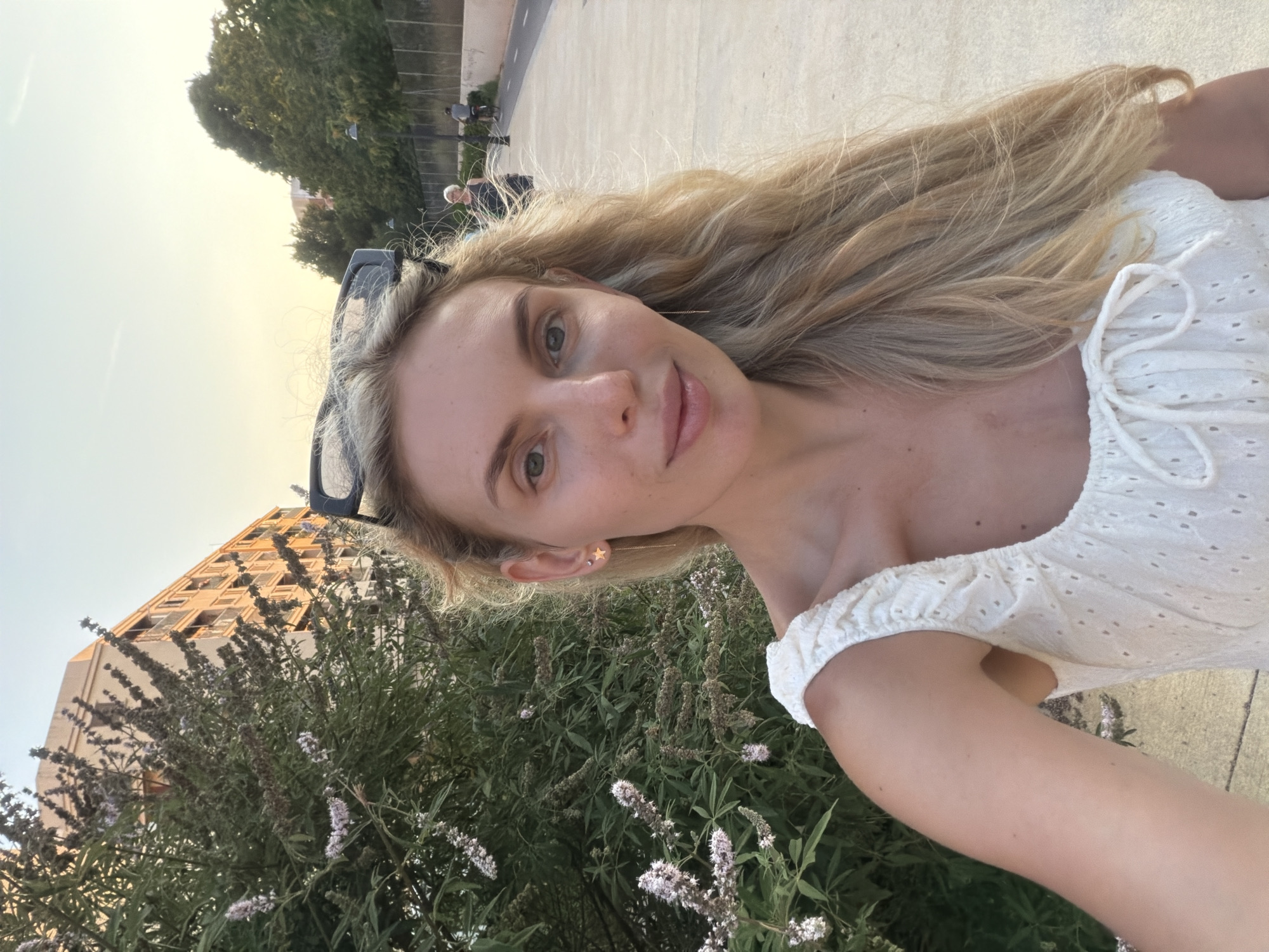 This is a portrait-oriented, full-body, outdoor selfie taken by a young Caucasian woman with long, blonde, wavy hair and light green eyes. She is wearing a white, off-the-shoulder top with eyelet detailing and a drawstring closure at the bust. A subtle star earring is visible in her left ear. She is smiling and looking directly at the camera, with a natural and pleasant expression.
Behind her, in the foreground, is lush green foliage with delicate purple flowers. Further back, to her left, a tall, multi-story building with visible windows and balconies is partially obscured by the greenery. To her right, a paved walkway can be seen, with several figures walking in the distance.
The lighting suggests it is late afternoon or early evening, with a warm, soft glow illuminating the scene. The sky is a pale yellow, indicating a sunset or sunrise. The overall atmosphere is relaxed and casual, possibly indicating a leisurely stroll or a moment captured during an outing. The location is identified as Benimàmet-Beniferri, Spain. No text is visible in the image.