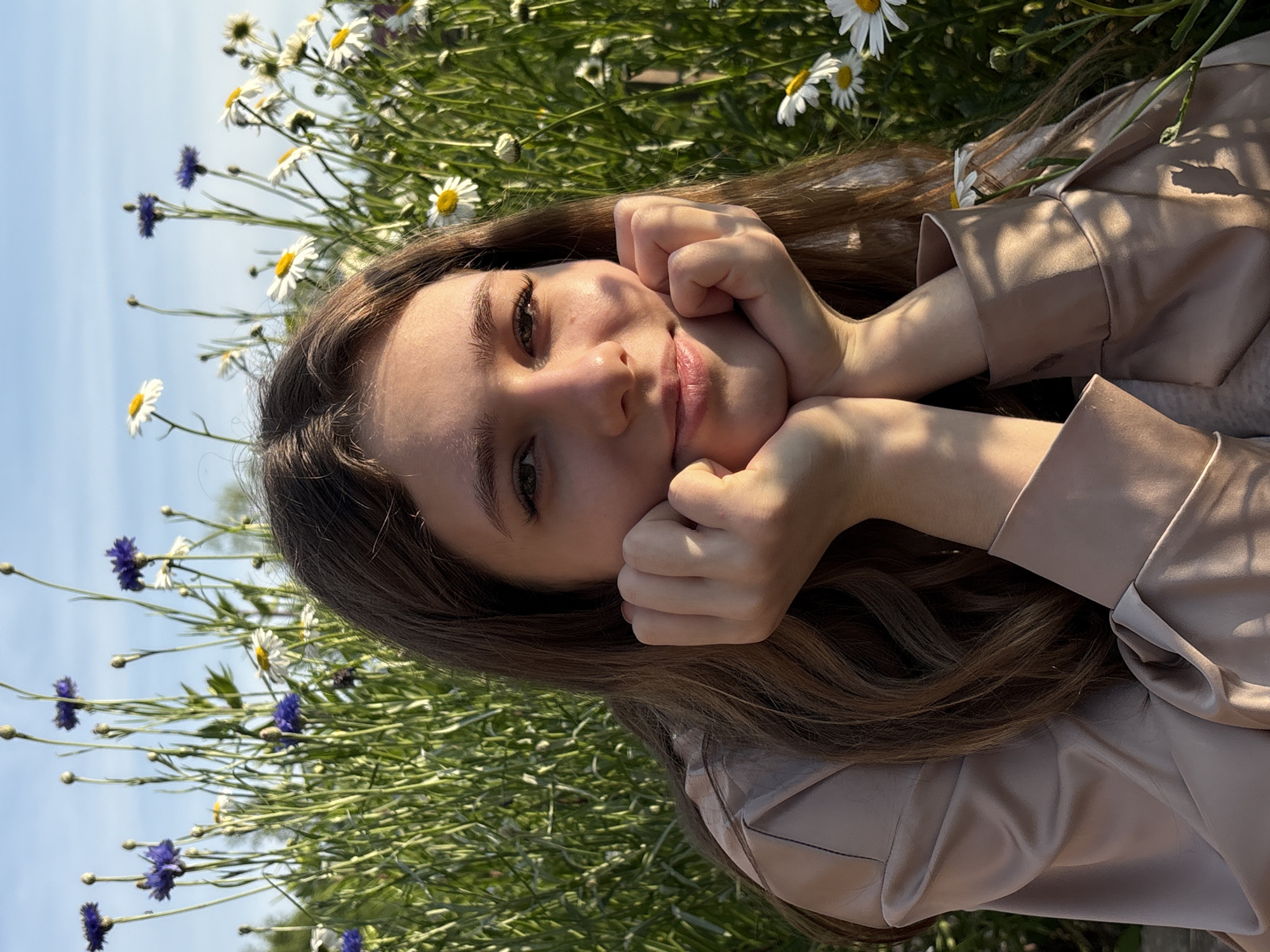 A young woman with long brown hair rests her chin on her hands, which are clasped together. She is looking directly at the camera with a gentle smile and her eyes are open. She is wearing a light-colored satin shirt. The background is filled with lush green grass and a variety of flowers, including white daisies and blue cornflowers. The sky above is a clear, soft blue. The lighting suggests it is late afternoon, with warm sunlight casting shadows on the woman and the foliage. The scene appears to be outdoors, possibly in a meadow or garden, in the city of Razdolnoye, Russia.