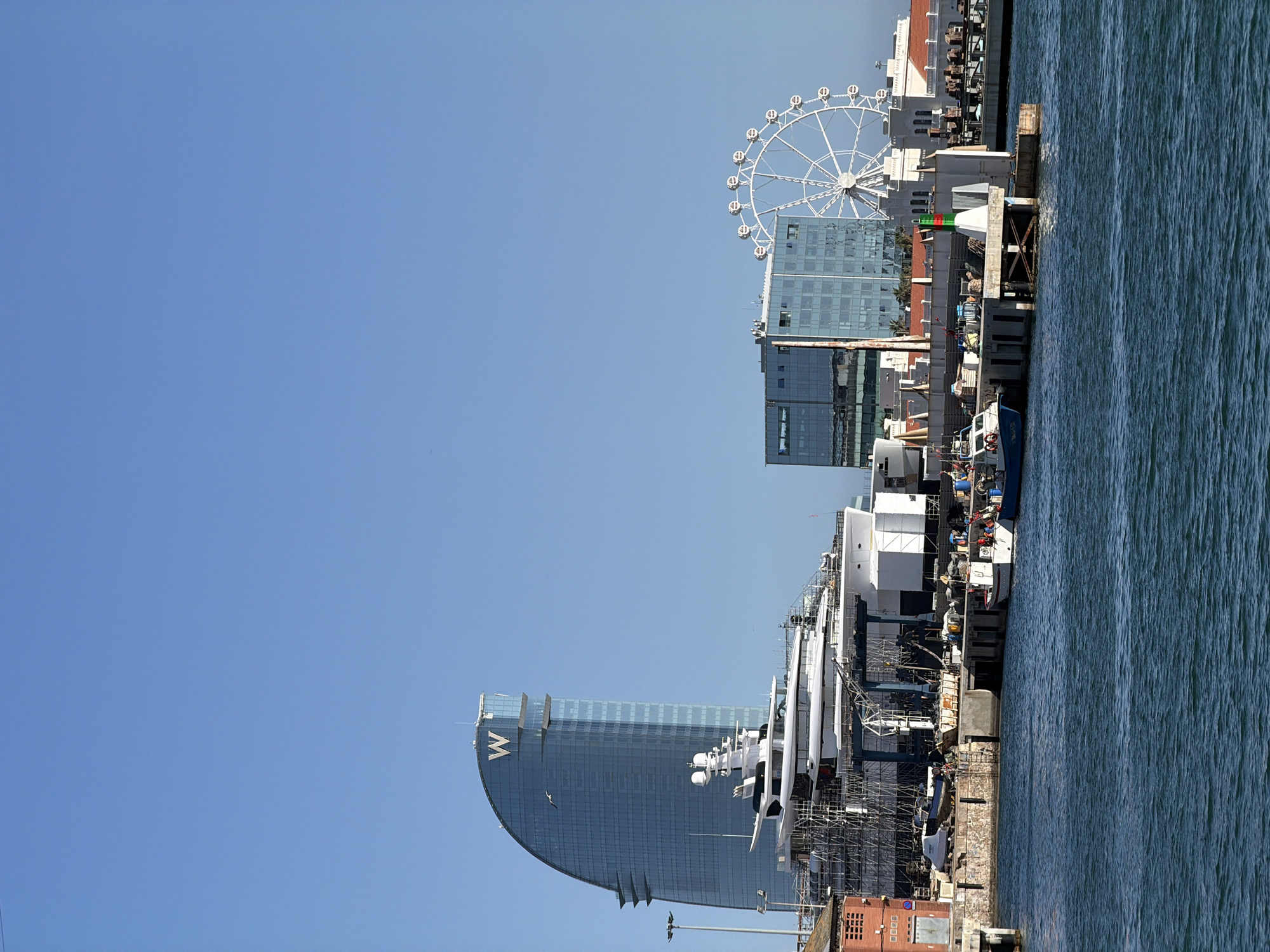 In Barcelona, Spain, a waterfront scene unfolds under a clear blue sky. Dominating the left side is the distinctive sail-shaped W Barcelona hotel, its glass facade reflecting the sunlight. In front of the hotel, a large white superyacht is undergoing construction or refurbishment, surrounded by scaffolding. To the right of the hotel, a modern glass-fronted office building stands, and further right, a white Ferris wheel is visible. The foreground features the blue water of the port, with several small boats docked along a pier. The pier is lined with various structures, including a brick building on the far left, storage containers, and utility equipment. A navigation buoy with red and green lights is positioned on a concrete structure extending into the water. The overall impression is one of urban development and maritime activity in a coastal city.