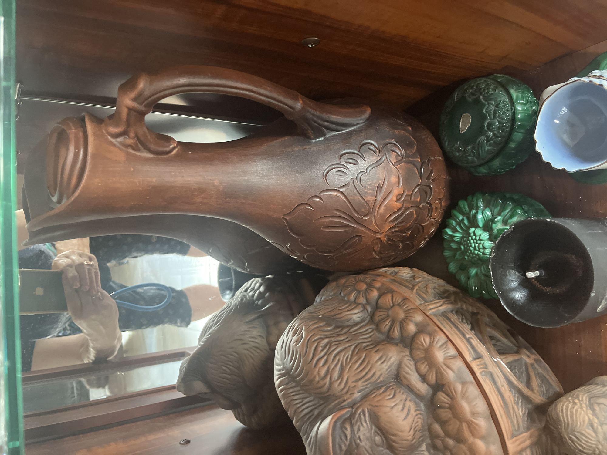 A wooden cabinet contains various decorative items. In the center is a large, brown ceramic pitcher with intricate floral designs and a sculpted handle. To the left of the pitcher is a ceramic lidded bowl shaped like a coiled animal, possibly a cat or rabbit, with floral motifs. To the right of the pitcher are two small, green ceramic decorative boxes, also with embossed designs. In the foreground, a black candle in a gray holder is visible, and to the right of that, a portion of a light blue ceramic cup is seen. The background reflects a person holding a smartphone, presumably taking the photo, and a tiled floor. The scene appears to be indoors, in what looks like a display cabinet.