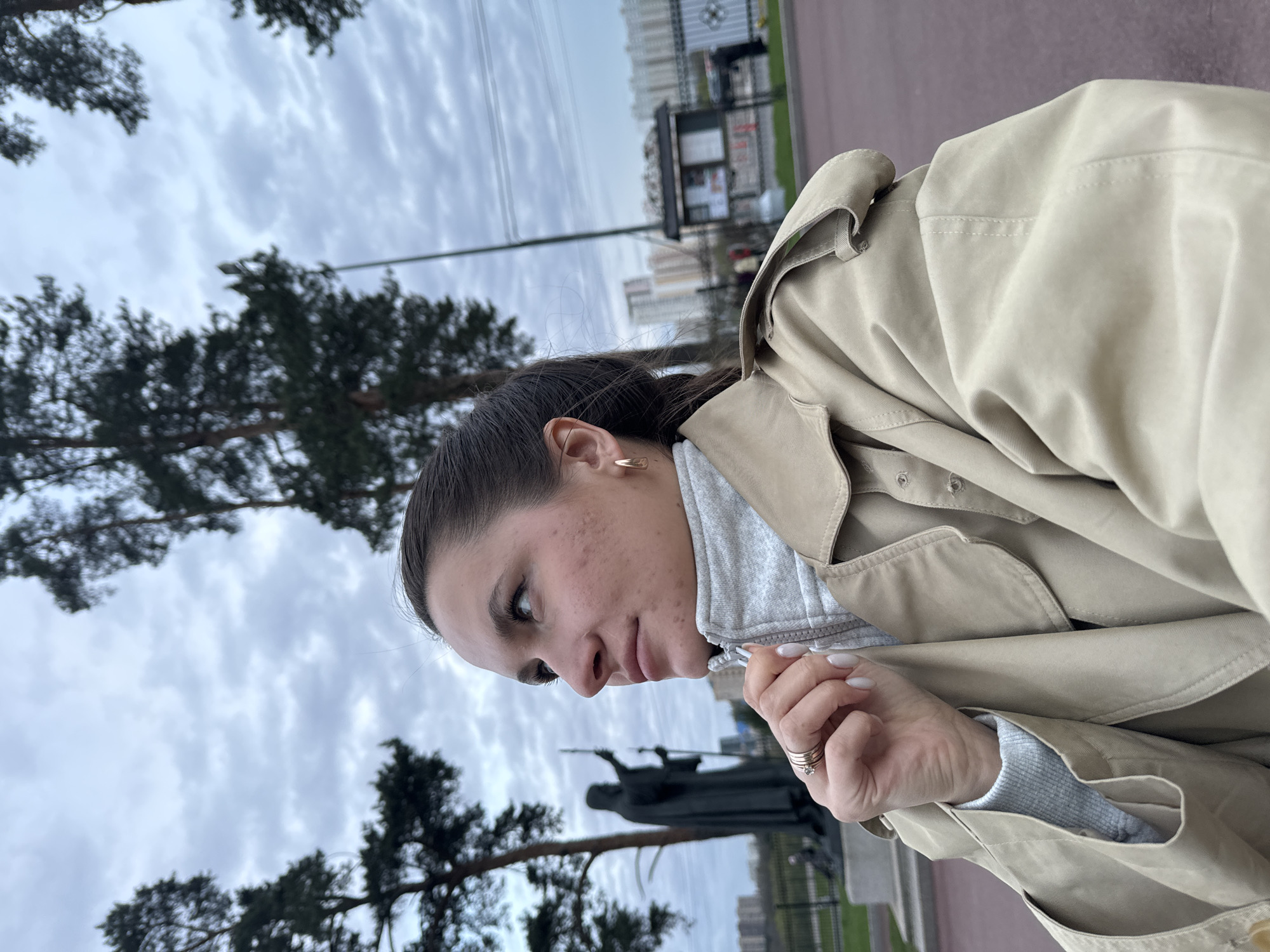 A woman stands outdoors in front of the "Monument to Children – Victims of Adult Vices" in Moscow, Russia. The sky is overcast. She wears a beige trench coat over a grey hooded sweatshirt. Her hair is pulled back, and she has an earring on her left ear. Her fingernails are painted a light color, and she wears rings on her right hand. In the background are dark green trees and a bus stop, with buildings visible in the distance. The statue is of a dark figure holding a scythe.