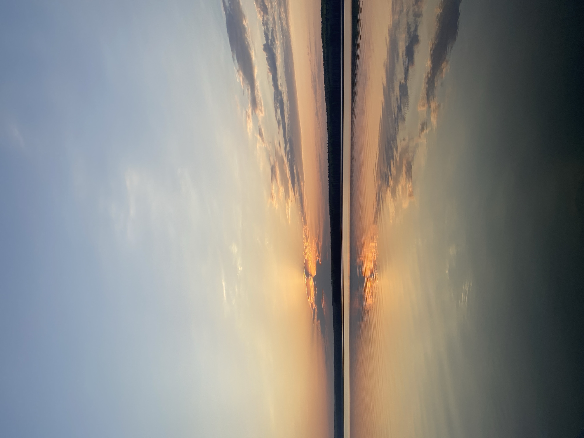 The image depicts a sunset over water, likely a lake or river. The sky is a gradient from blue to orange with scattered clouds reflecting the light. The line between the sky and water is sharp, with a dense strip of trees visible in the middle ground. The water reflects the sky, creating a symmetrical effect. It is likely taken in Chaikovsky, Russia, considering the request.