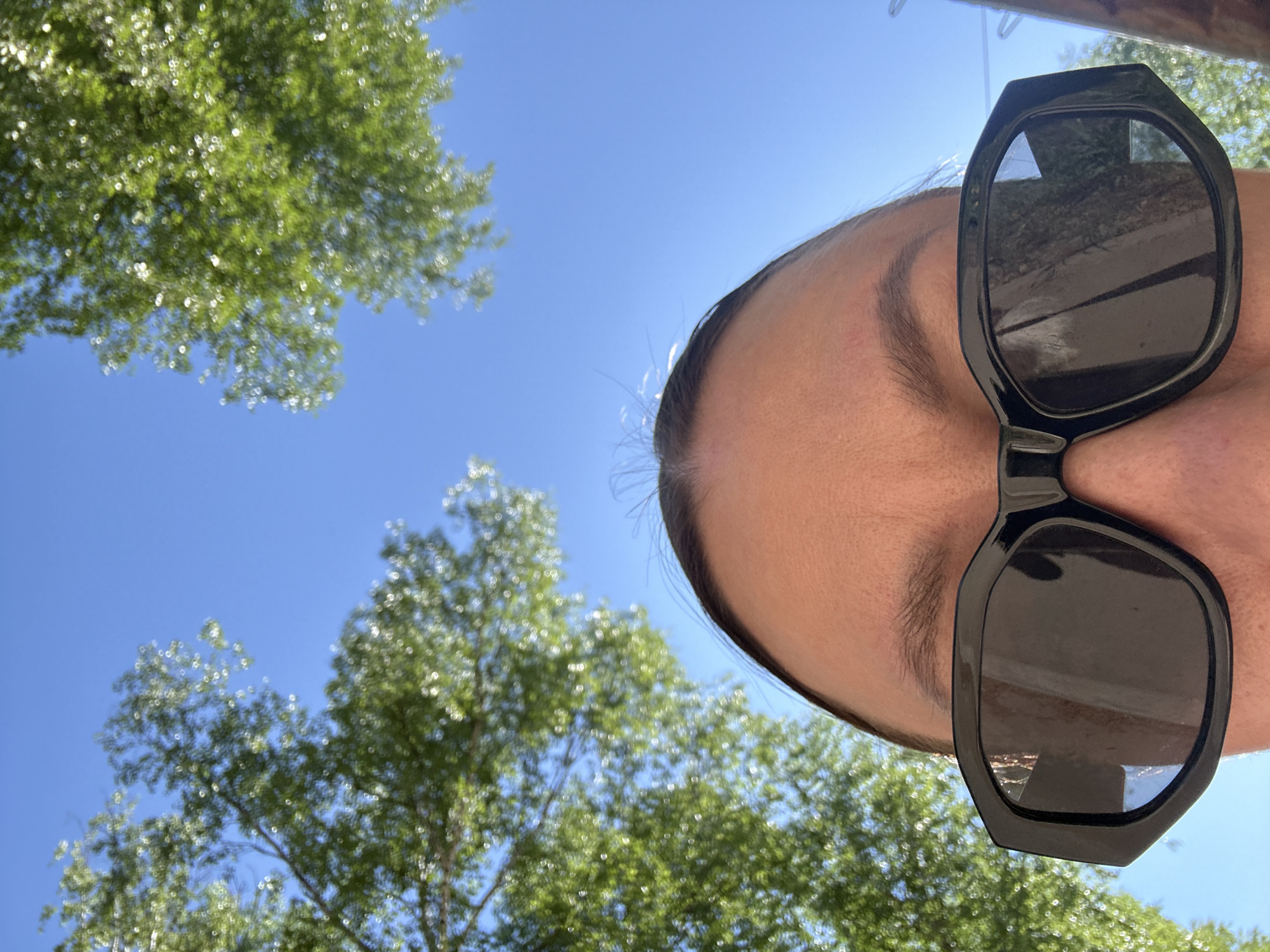 The image shows a person from a low-angle perspective, likely a selfie taken while looking upwards. The person is wearing large, dark sunglasses. Part of the person's forehead, eyebrow, and hair are visible. The background features a bright blue sky and green tree foliage, suggesting an outdoor location. There is no information available about the location being in the city of Votkinsk, Russia.
