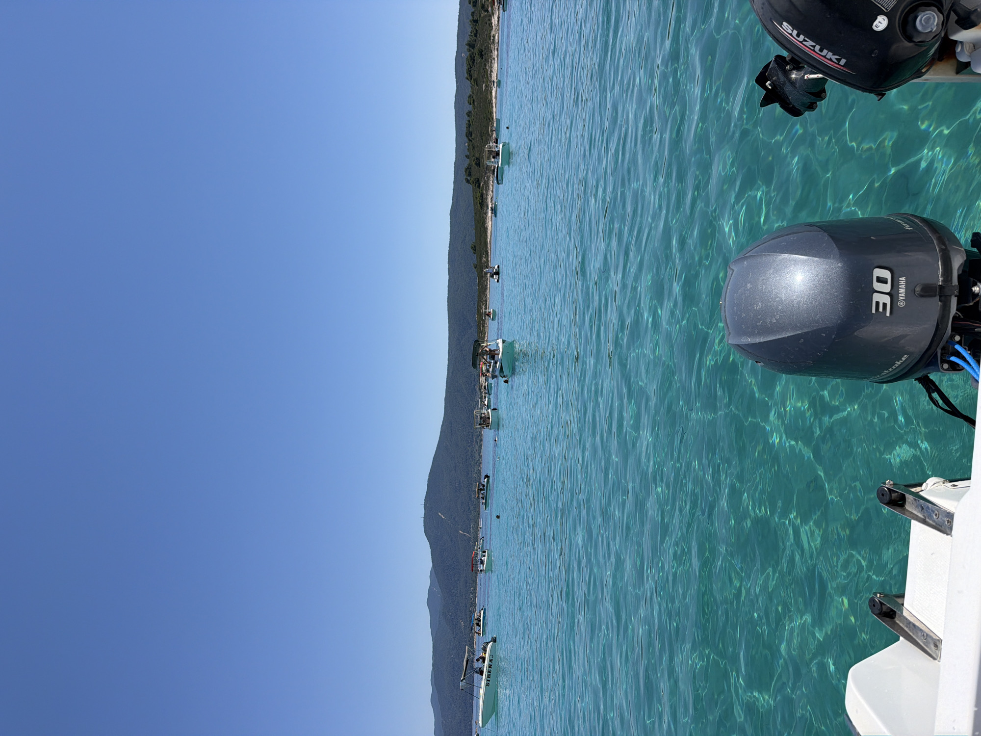 The image shows a view from a boat on a clear, turquoise sea under a bright blue sky. In the distance, a line of boats is anchored or moored near a wooded coastline with hills behind it. The foreground of the image features the back of the boat the photographer is on, with two outboard motors visible. One motor is a dark grey Yamaha with "30" prominently displayed, and the other is a black Suzuki. The water is exceptionally clear, allowing the rippled seabed to be seen beneath the surface. There are no visible signs of a city or urban environment; it appears to be a coastal or island scene in Greece.