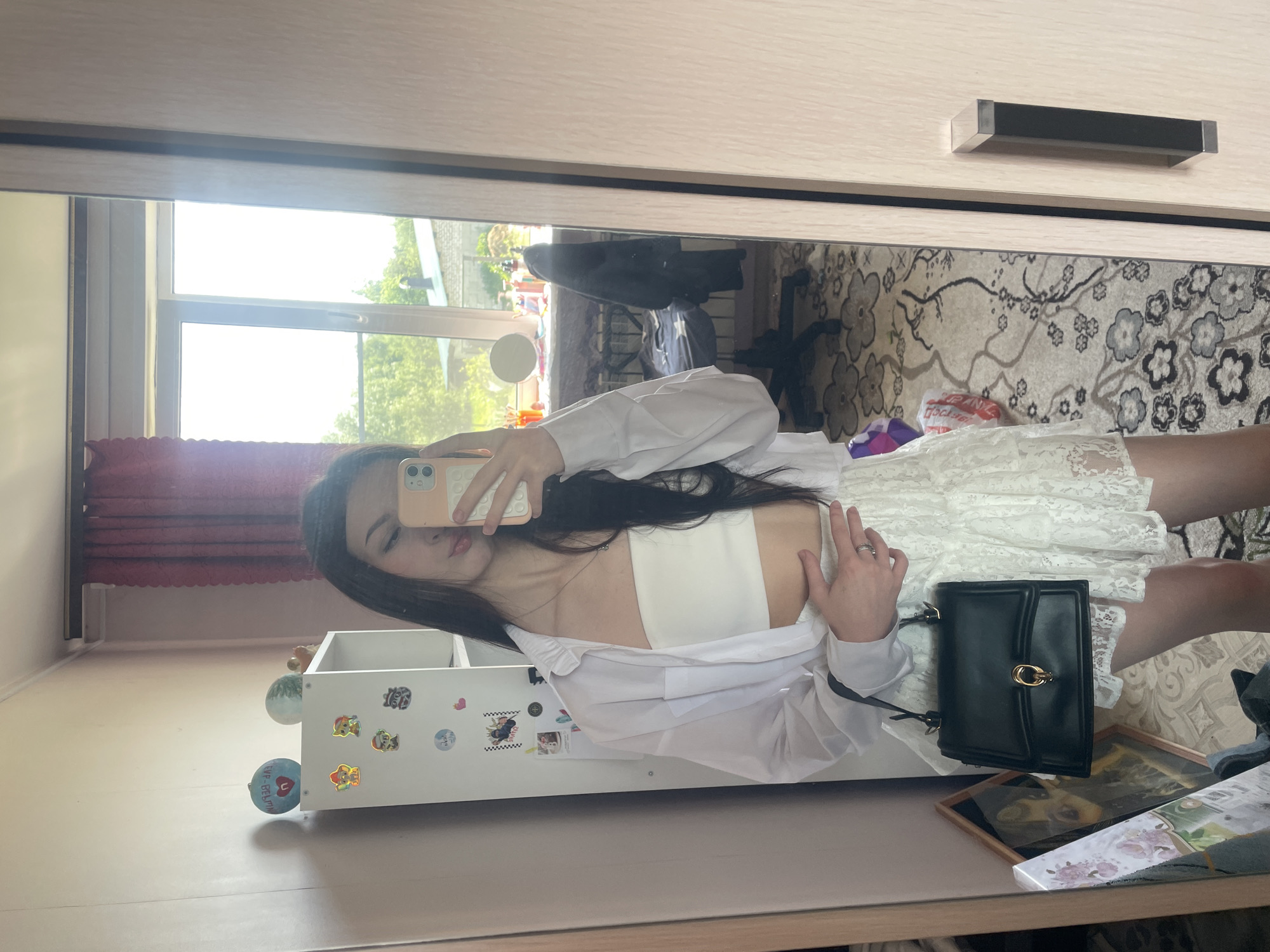 A young woman with long, dark hair takes a mirror selfie in a room. She is wearing a white bandeau top, a white unbuttoned shirt, and a white lace skirt. She is holding a black handbag with a gold clasp. In the background, a window shows trees and buildings. A closet door is visible to the right of the mirror. The floor is covered with a patterned rug. The overall setting appears to be a residential interior.