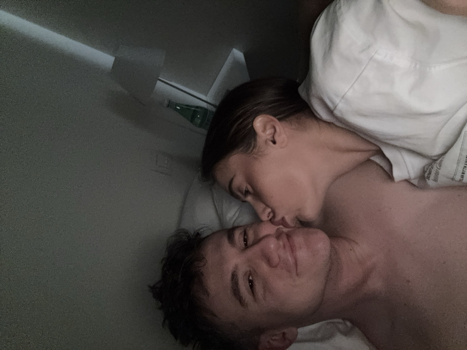 The image presents two individuals, a male and a female, in a dimly lit indoor setting, consistent with a bedroom in Venice, Italy. The male, with dark, disheveled hair, lies on his back, looking towards the camera with a subtle smile. He is bare-chested and partially covered by white bedding. The female, with dark hair, is positioned to his right, leaning in to kiss his left cheek/mouth area. She is wearing a light-colored t-shirt. In the background, to the right, a white wall features a wall-mounted bedside lamp with a white shade. Below the lamp, on a small shelf or surface, a green glass bottle, consistent with a water bottle, is visible alongside a wall outlet or switch.