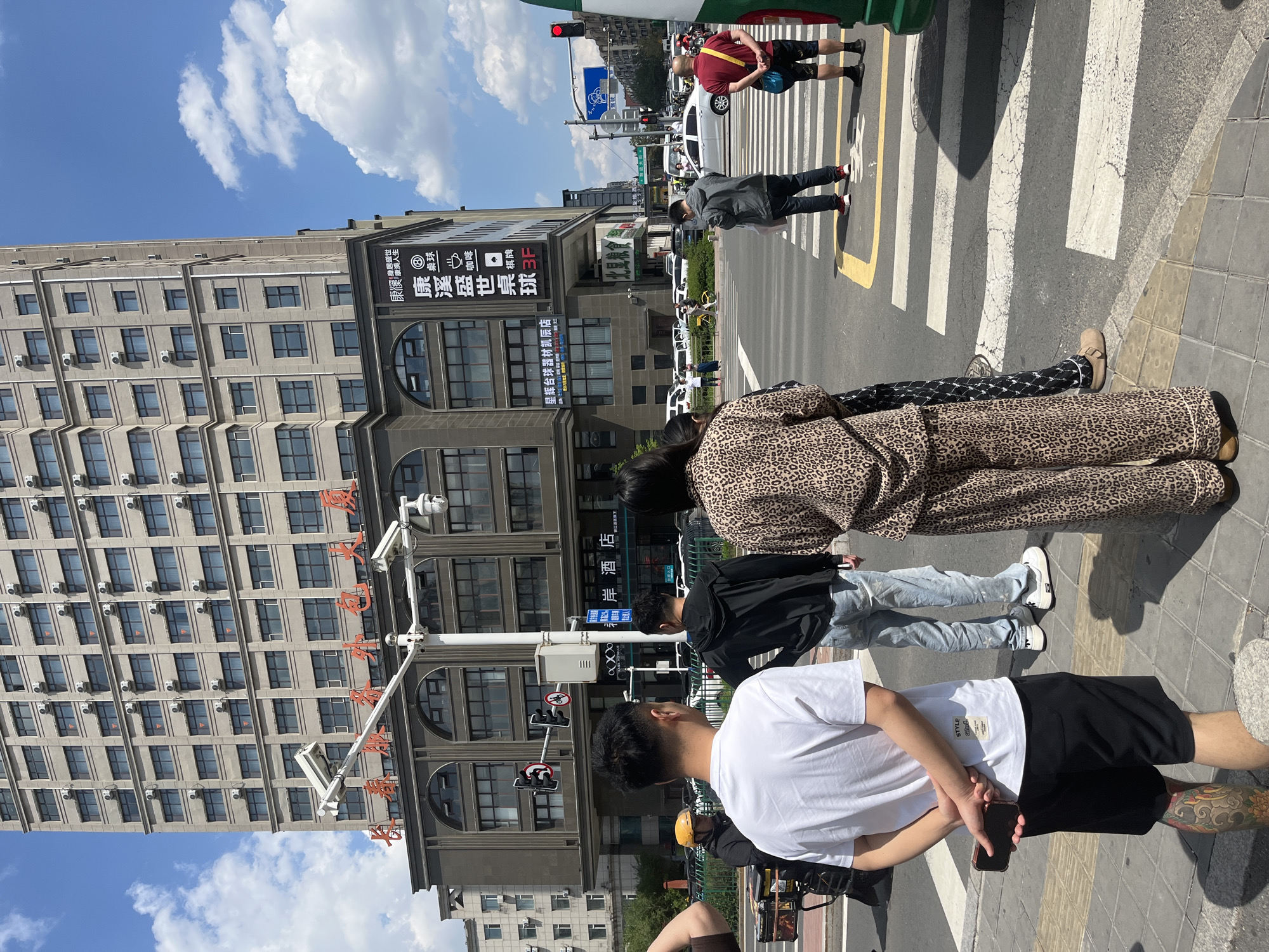 This is a street scene in Changchun City, China. A group of pedestrians is standing at a crosswalk on a sunny day. The foreground includes a man in a white shirt and black shorts with tattoos on his leg holding a phone. Behind him is a person in a black jacket and blue jeans, and a woman in an animal print outfit. In the background, people are crossing the street. A tall, grey building with multiple rows of windows dominates the left side of the frame. Chinese characters are displayed on the building's facade. A traffic light is visible above the crosswalk.