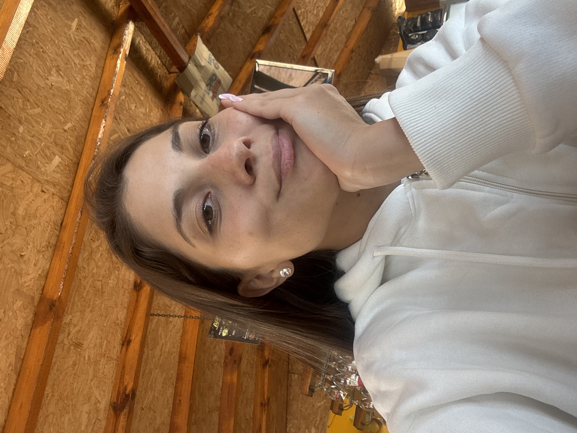 A woman is posing for a selfie, looking up at the camera, with her hand resting on her face. She is wearing a white hoodie and a stud earring. The background appears to be a partially finished or unfinished room with exposed wooden framing and wood paneling. Given the reference to Sozopol, Bulgaria, the location is likely inside a home or building, but no specific landmarks or contextual clues in the image directly confirm the city. The woman's interaction is with the camera, suggesting she is the subject of the photograph.