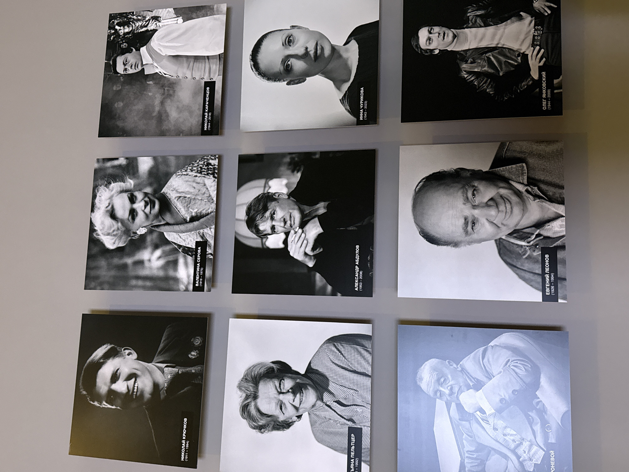 The image shows a collection of nine black and white photographs displayed on a light-colored wall. Each photograph is a portrait of a different person, with a caption below in Russian, presumably identifying the individual and their birth and death years. The portraits are arranged in a 3x3 grid. Based on the captions, the people in the photos are actors and actresses. From left to right and top to bottom, they are: Nikolai Kryuchkov, Tatyana Peltzer, Evgeny Leonov, Valentina Serova, Nikolai Karachentsov, Alexander Abdulov, Inna Churikova, and Oleg Yankovsky. The wall appears to be indoors. The location is in Moscow, Russia.