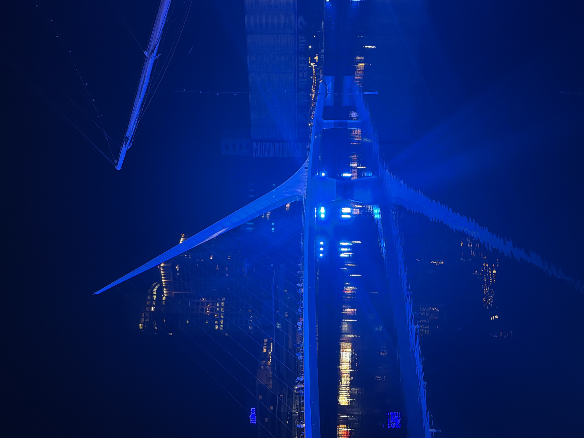 The image shows a night scene of the Puente de la Mujer (Woman's Bridge) in Buenos Aires, Argentina. The bridge and surrounding buildings are illuminated with blue light, which is also reflected in the water beneath the bridge. The bridge is a modern pedestrian bridge with a single mast and cables supporting a rotating section. The reflection of the city's lights and buildings is visible in the water.