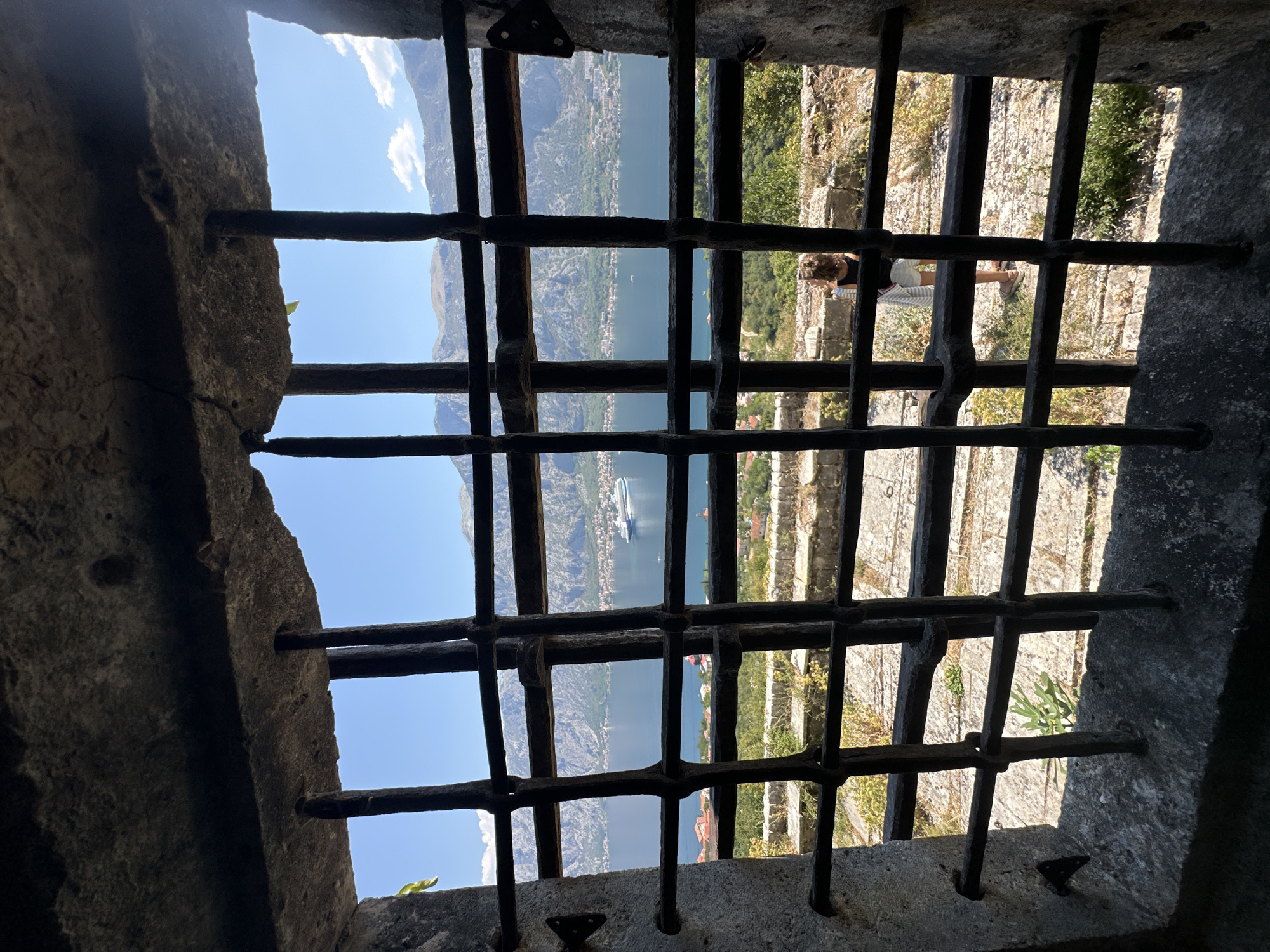 The image depicts a view through a barred window of a stone structure, likely part of a historic fortification, in Prčanj, Montenegro. The bars are thick and dark, set into a rough stone frame. Through the bars, a panoramic vista unfolds. A large, white cruise ship is visible on the calm, blue water of a bay, likely the Bay of Kotor. The bay is surrounded by rugged, green mountains that rise steeply from the shoreline. Along the coast, houses and buildings of a town are clustered. In the foreground, outside the window, a young person with curly brown hair, wearing a white and gray striped skirt and a dark top, stands with their back to the viewer, looking out at the scenery. They are standing on a stone-paved area with some sparse vegetation. The lighting suggests a bright, sunny day.