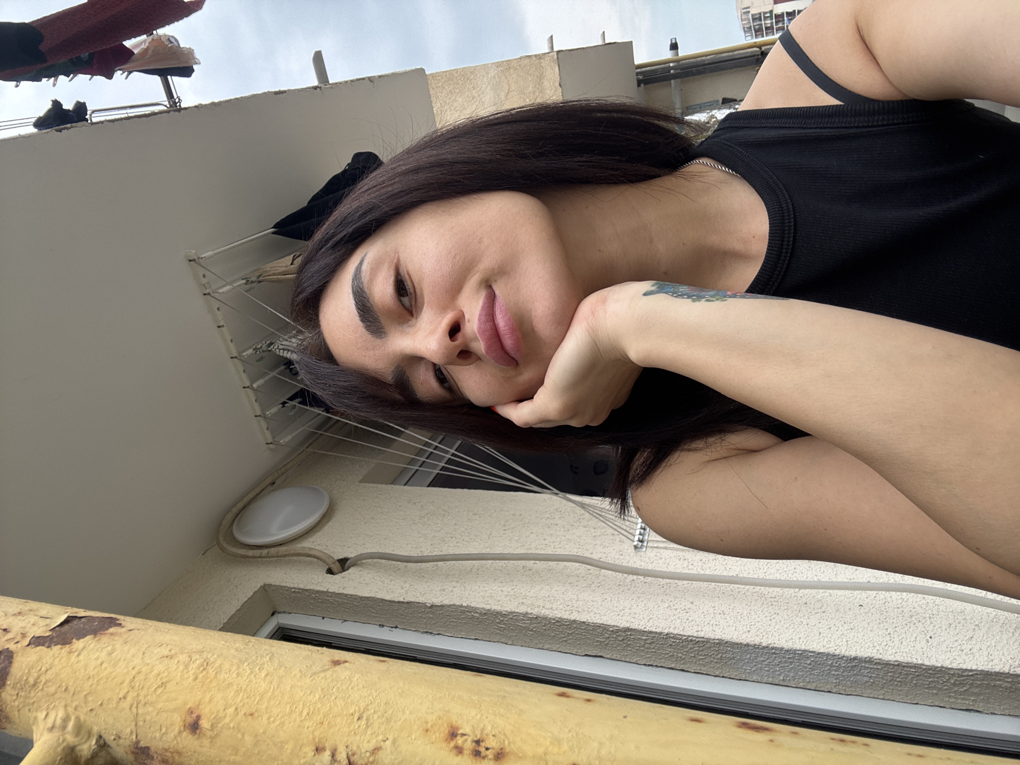 A selfie taken from a low angle shows a woman with dark hair and light skin, resting her chin on her hand. She is wearing a black tank top and has a tattoo on her arm. Behind her, a building with a textured beige facade and a yellow pipe are visible. Clothes hang on a drying rack on a balcony overhead, and the sky appears overcast. The scene is likely an outdoor residential setting, possibly a balcony, in Tbilisi, Georgia, judging by the architectural style and common elements like external pipes and balconies with clotheslines.