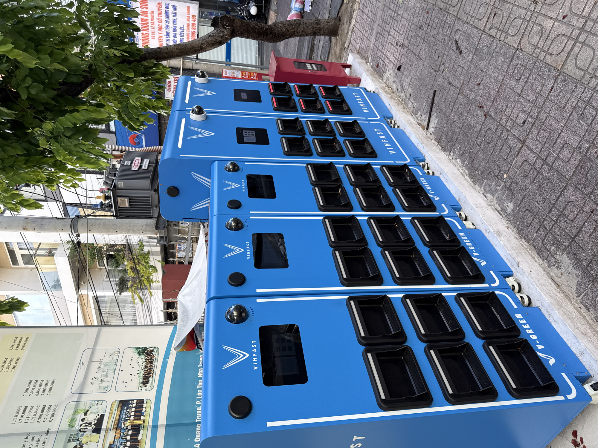 The image shows a row of bright blue battery swapping stations for VinFast electric scooters, located on a sidewalk in Nha Trang, Vietnam. The stations have the VinFast logo prominently displayed and are equipped with numerous compartments for battery storage. Two security cameras are visible mounted on the top of the stations. Adjacent to the stations, there is a red fire alarm box and a tree with lush green leaves. In the background, a transformer and several buildings are visible. The sidewalk is paved with patterned tiles, and some debris is scattered on the ground. A street sign in the background indicates the location as "Quang Trung, P. Lộc Thọ, Nha Trang."