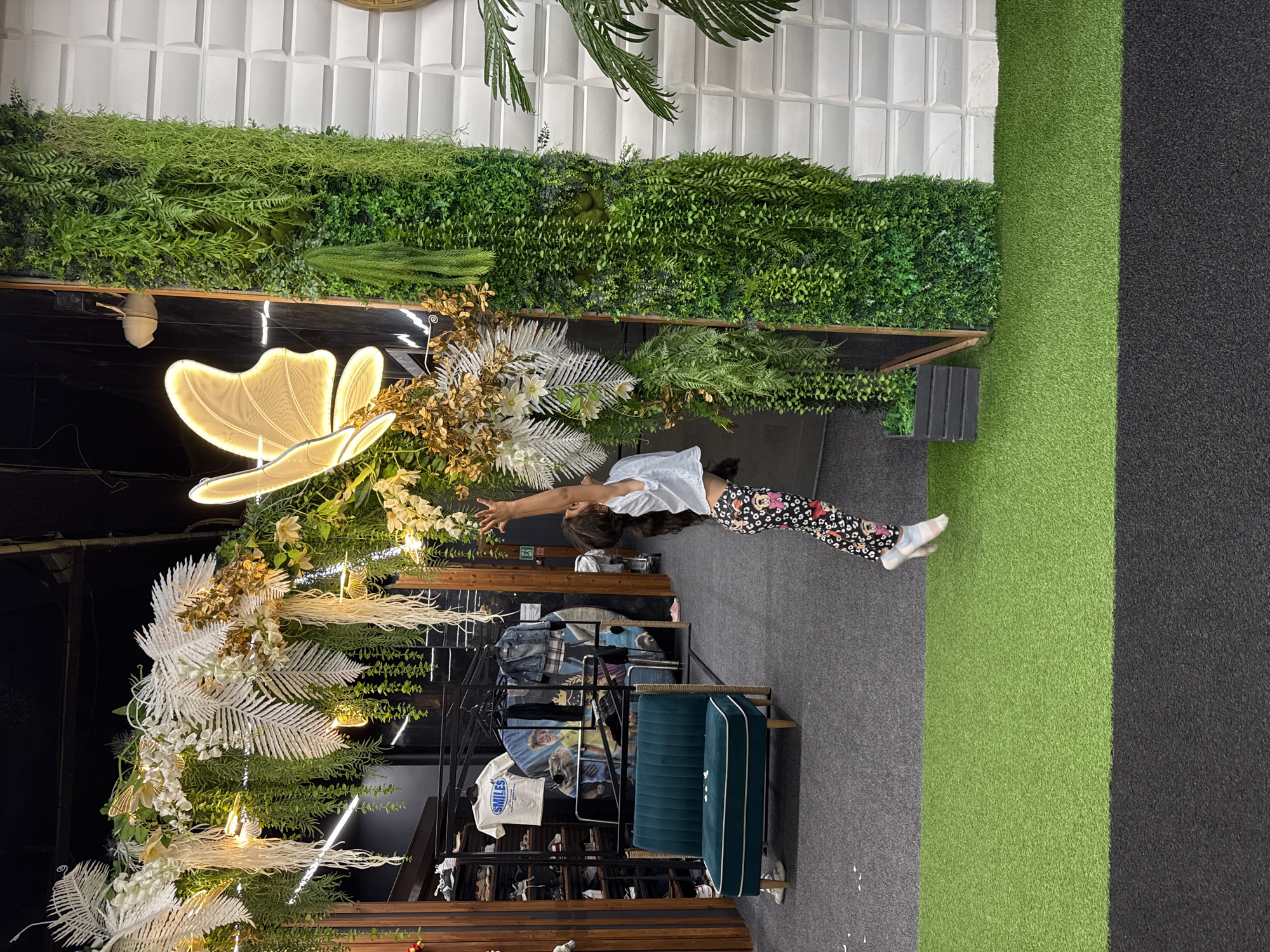 A young girl with dark hair, wearing a white top and patterned leggings, is reaching upwards towards a large, illuminated butterfly decoration. She is in mid-jump, with her right leg extended and her left foot on the ground. The girl is positioned on a green carpeted area. Behind her, the environment appears to be a retail space or an exhibition, with shelves of clothing and shoes visible in the background. There is a teal-colored bench with white piping. A large, vertical wall covered in lush green artificial foliage is to the right of the girl, and next to that is a white textured wall. The scene is indoors and well-lit. This image was captured in Sochi, Russia.