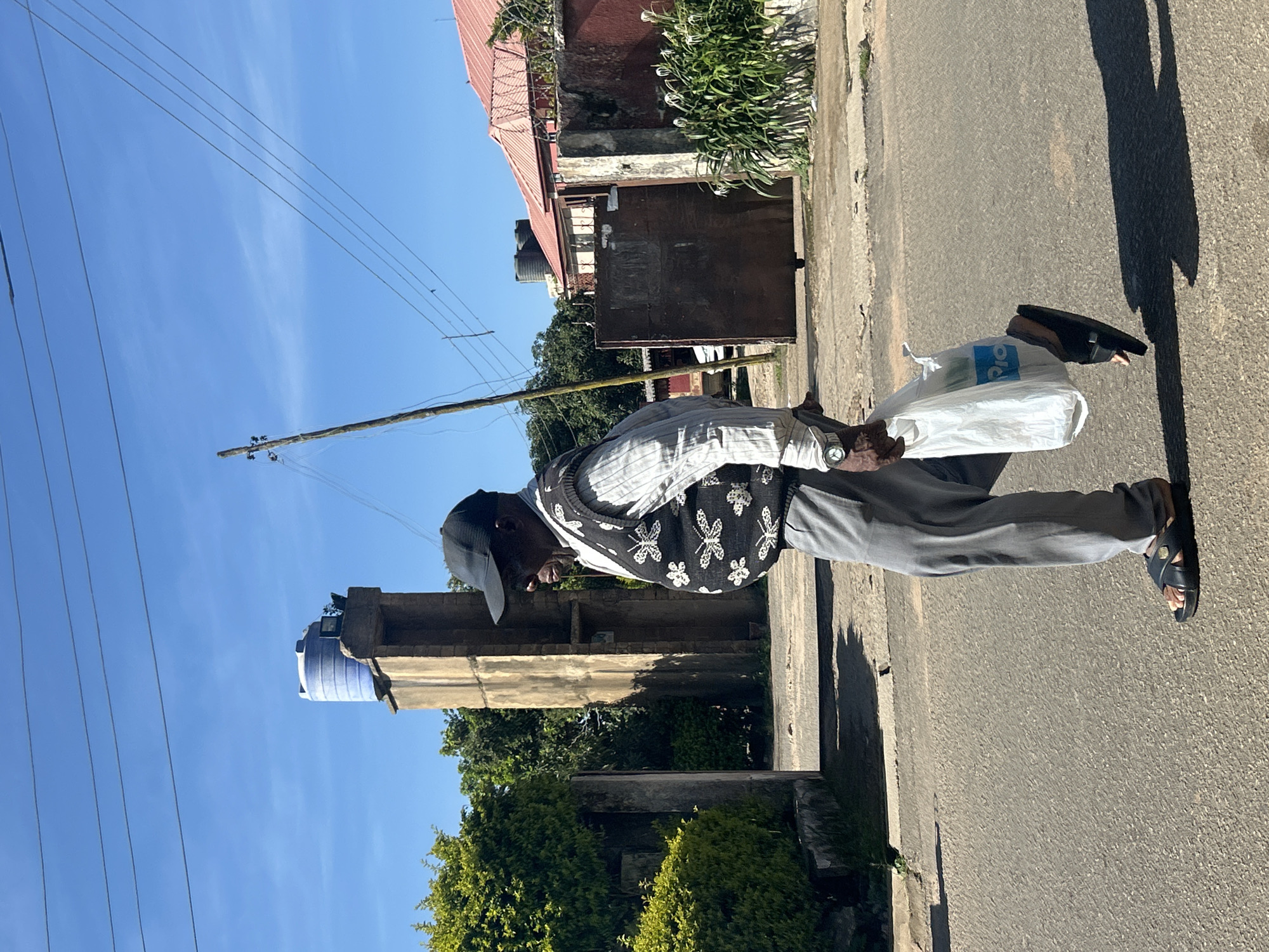 An elderly Black man wearing a black baseball cap, a light-colored pinstriped shirt, and a black vest with a white butterfly pattern walks on an asphalt road, carrying a white plastic shopping bag in his right hand. He is wearing grey trousers and black sandals. To the left of the man, there is a gatepost and dense green foliage. Behind the man, a concrete structure with a blue water tank on top stands tall against a clear blue sky. The background also shows a building with a red corrugated roof and a large metal gate. The scene appears to be a street in Jos, Nigeria, under bright sunlight, casting a distinct shadow of the man onto the road.