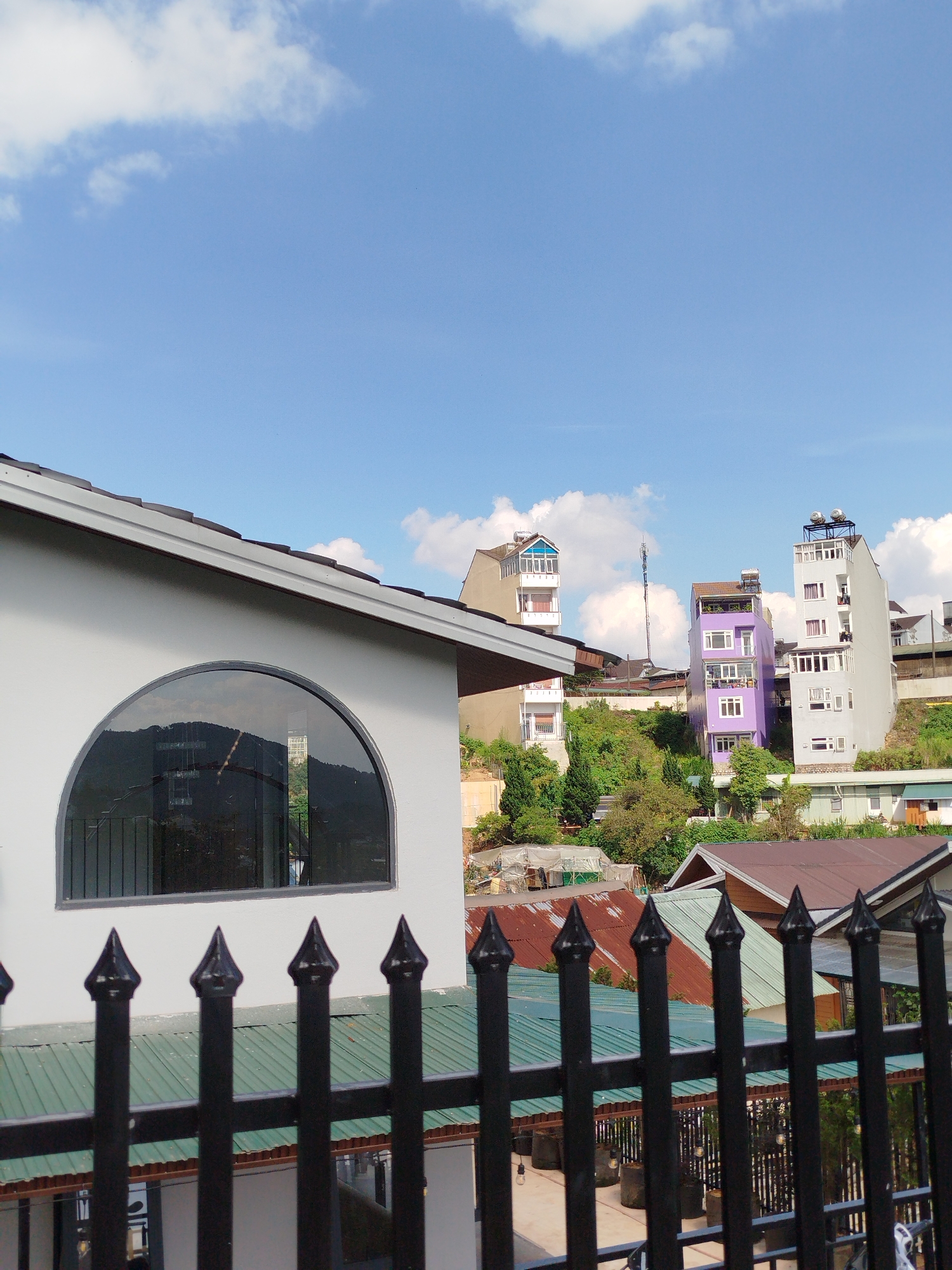 The image captures a cityscape in Da Lat, Vietnam. A foreground black metal fence with pointed posts frames the view. Behind the fence, a light gray building with a distinctive arched window is partially visible. The window reflects a mountainous landscape, suggesting a high vantage point. Beyond the gray building, the landscape consists of a collection of buildings of varying heights, colors, and architectural styles nestled on a hillside. Among them are buildings painted purple and white. Some buildings have rusty or green roofs. The roofs of other structures are visible beneath the fence line, indicating the city's tiered layout on the slopes. The background features a blue sky with scattered white clouds. The weather appears clear and sunny.