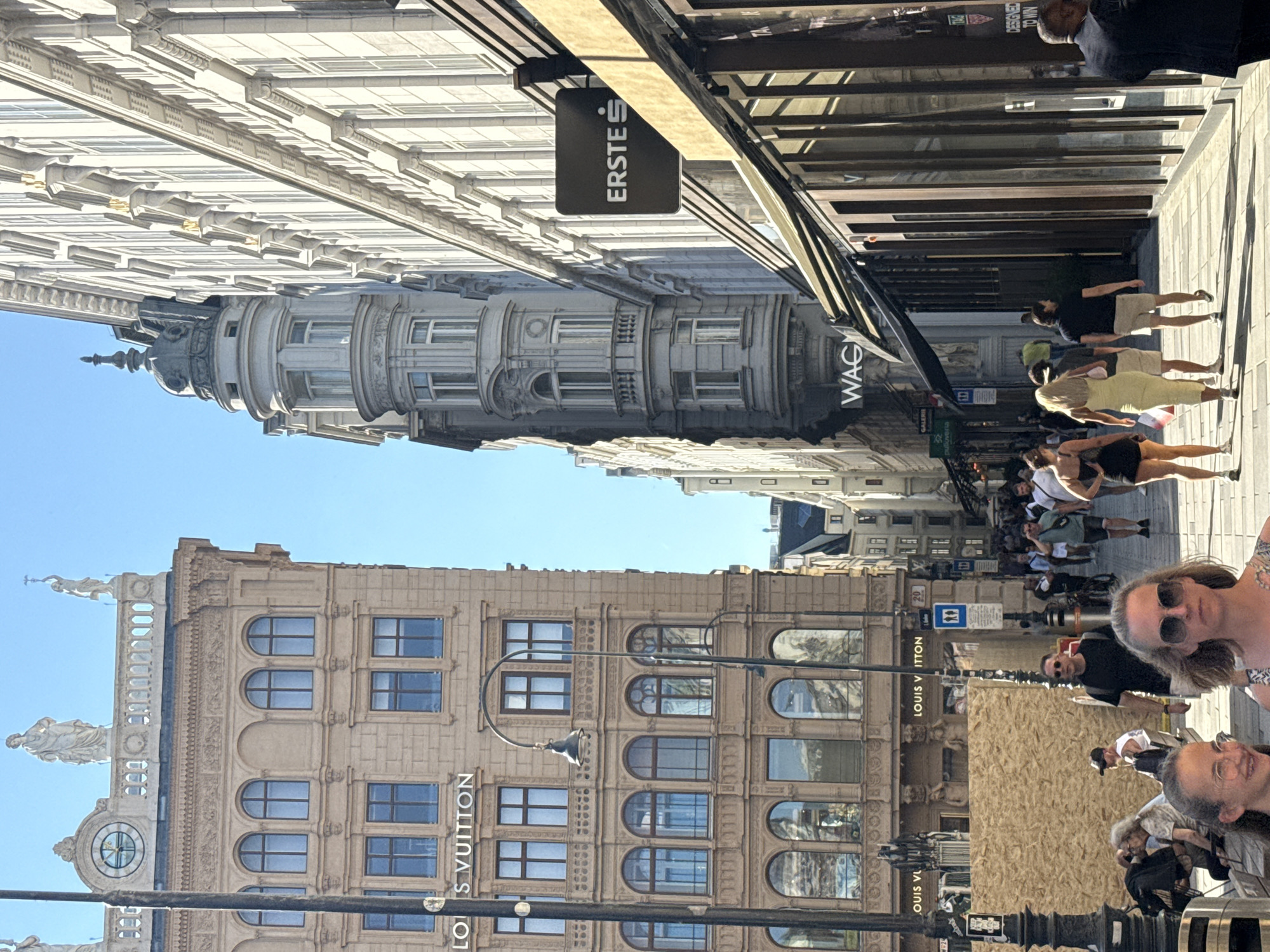 The image shows a bustling street scene in Vienna, Austria, specifically on Graben street, a popular shopping area. On the left, a large, ornate building with arched windows and statues on its roof is visible. The building features the "LOUIS VUITTON" signage. A lamppost stands in front of it. Across the street, another building with a distinctive, rounded architectural element and a dark "ERSTE$" sign is seen. People are walking along the pedestrian street, some are shopping, and others are simply passing through. The street is paved with cobblestones and lined with shops. The weather appears to be sunny with a clear blue sky.