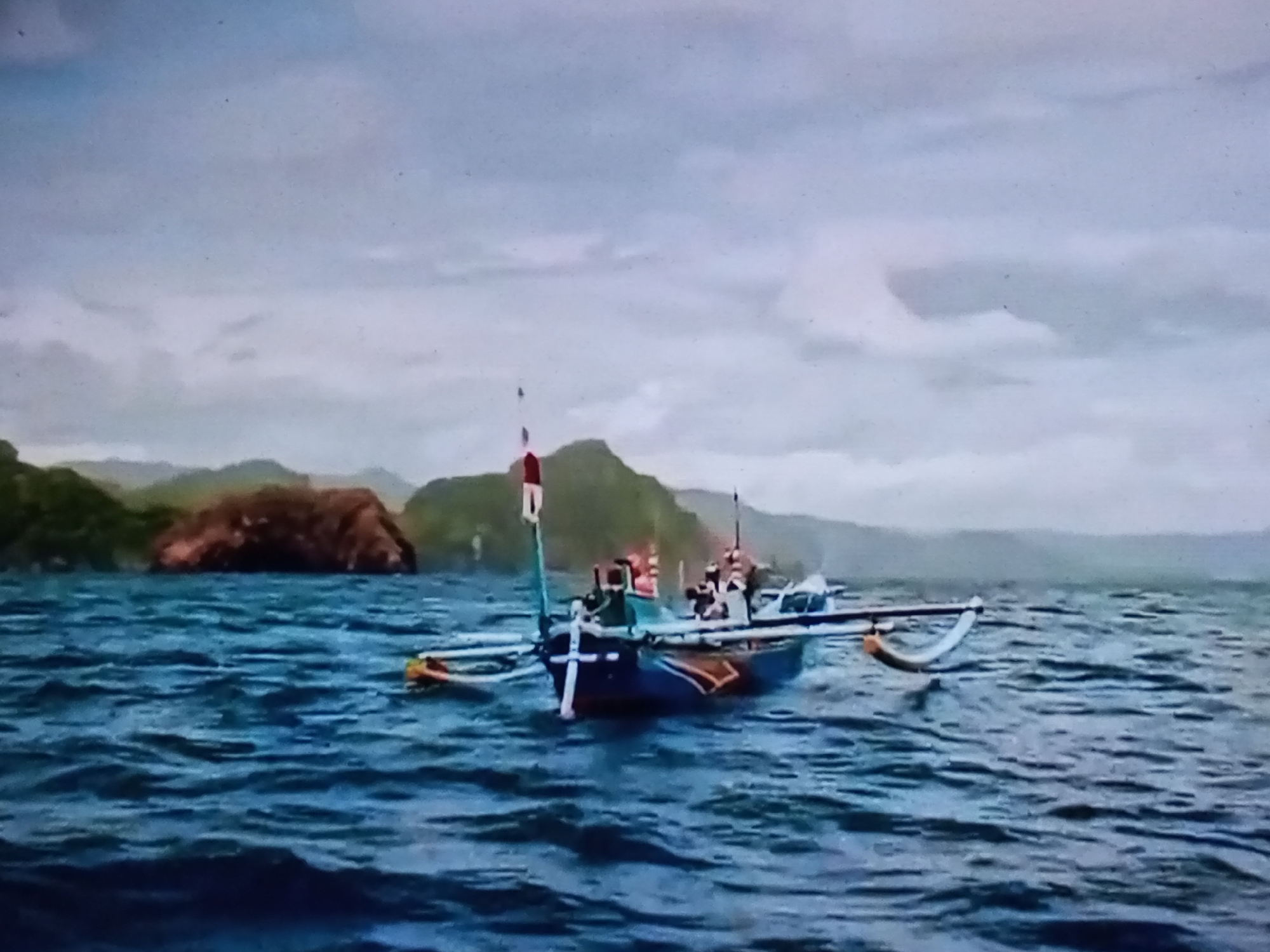 An overcast day on choppy, dark blue water with a traditional Indonesian fishing boat prominently featured in the foreground. The boat, painted with blue, red, and white accents, has a outrigger on the right side and a prominent mast with a red and white flag. Several people are visible on board the boat, some appearing to be in motion. In the background, a lush green, mountainous coastline with a large, reddish-brown rock formation is visible. The sky is filled with grey clouds. This scene is consistent with the coastal environment of Banyudono, Indonesia, known for its fishing villages and scenic coastlines.