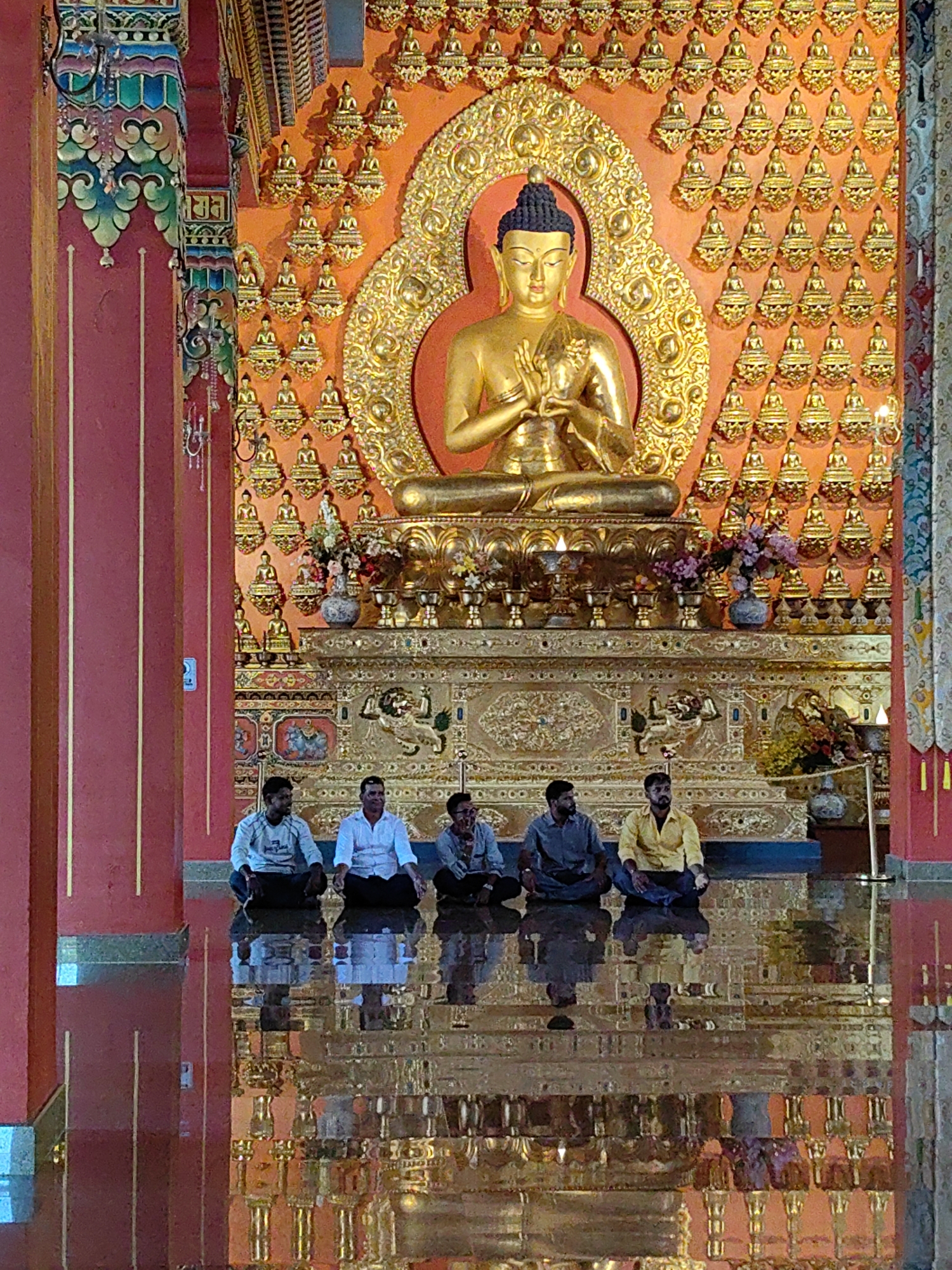 In Parsa, Nepal, inside a temple, five men are seated on the floor facing a large golden Buddha statue. The statue is centrally positioned against an orange wall adorned with numerous smaller golden Buddha figures. The floor is highly reflective, mirroring the intricate golden altar and the seated figures. The temple's interior features ornate golden decorations and vibrant red pillars. The men are dressed in casual clothing, appearing to be meditating or observing the statue.