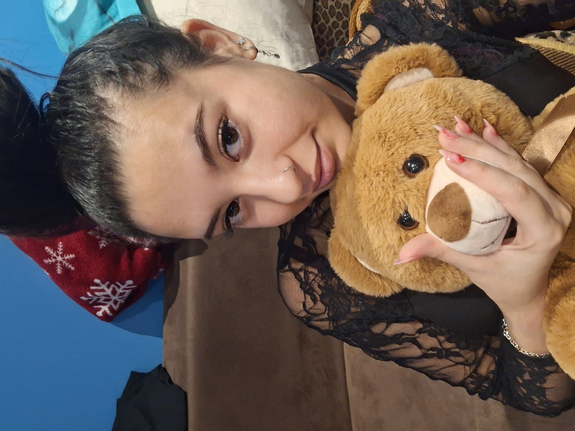 A young woman with dark hair pulled back in a bun poses indoors, lying on a beige or brown surface. She is wearing a black lace top and holds a brown teddy bear with a light-colored muzzle close to her face. Her left hand is visible holding the teddy bear's muzzle. She has a nose piercing and earrings in her left ear. In the background, there's a red cushion with white snowflake patterns on a blue surface, a white pillow, and a dark-colored garment. The image does not provide enough information to determine the specific location in Sofia, Bulgaria.