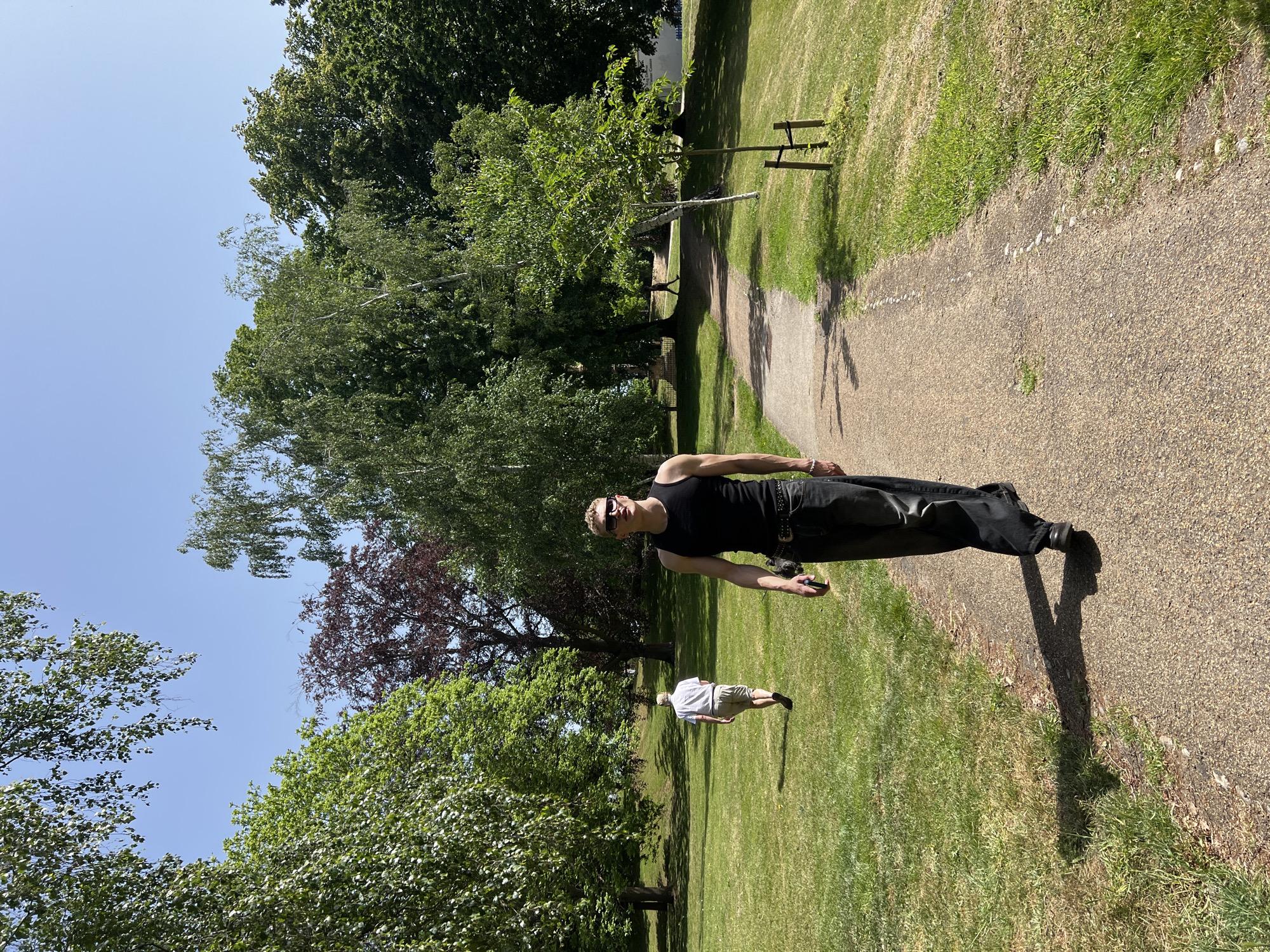 The image depicts a park scene, likely in Ipswich, United Kingdom, given the location provided. A light-skinned male is walking on a gravel path in the foreground; he wears a black tank top, dark pants, and sunglasses. He appears to be holding a dark-colored object, possibly a cell phone, in his right hand. His shadow extends onto the path and the adjacent grass. In the background, a second light-skinned individual, wearing a light-colored shirt and shorts, walks on the lawn parallel to the path. The environment consists of green grass, mature trees, and other shrubbery, suggesting a park or recreational area. A clear blue sky is visible through the branches of the trees. A small, newly planted tree is held in place with wooden stakes.