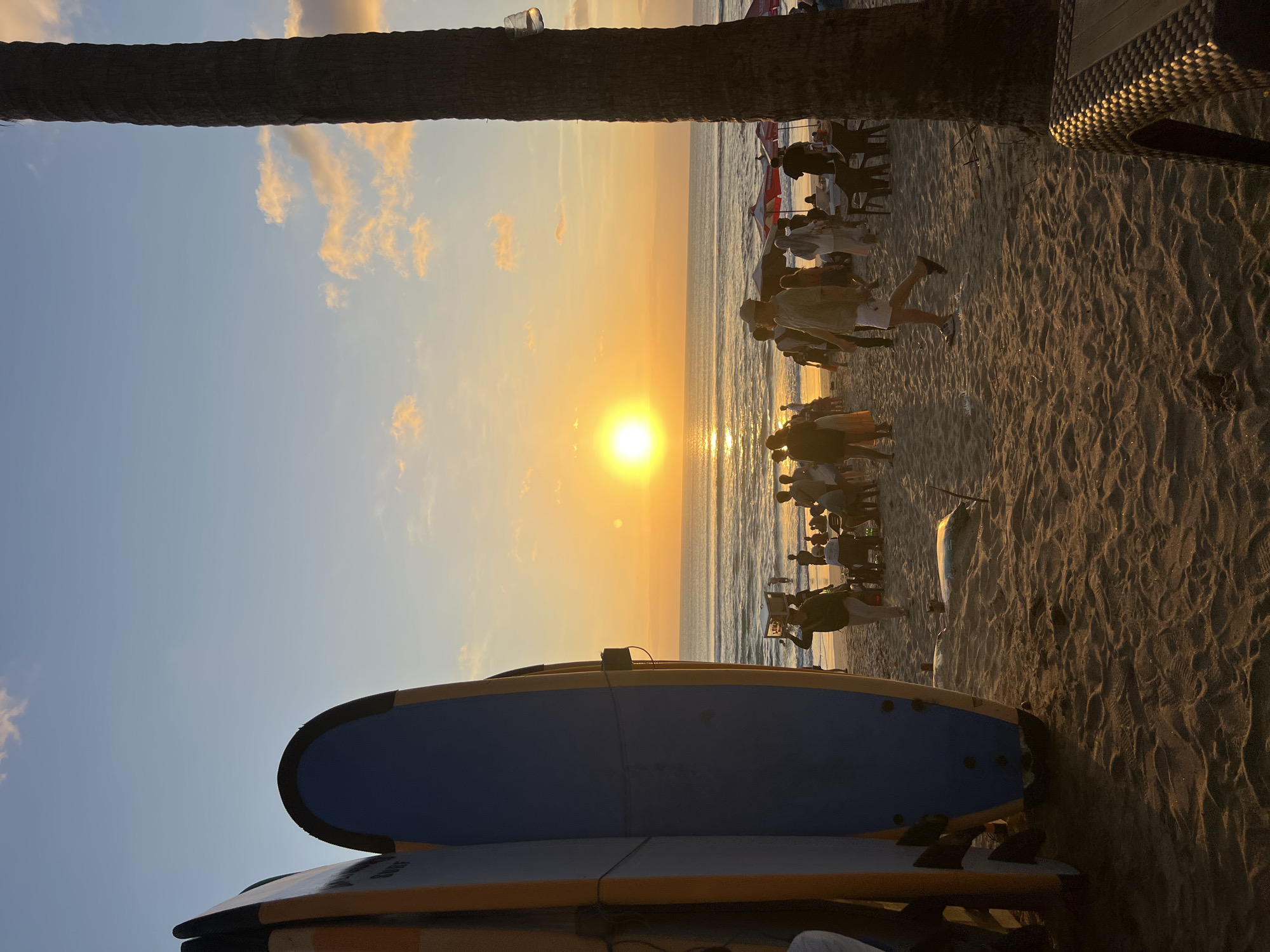 The image depicts a crowded beach scene in Kuta, Indonesia, during sunset. The sun is low on the horizon, casting a warm orange glow across the sky and water. Several surfboards are leaning against a palm tree in the foreground on the left. A group of people are gathered on the sand, some walking, some sitting, and some standing in conversation. In the background, more people are visible near the water's edge, and umbrellas suggest beachside service. The sand is covered in footprints. The overall atmosphere suggests a lively and popular tourist destination.