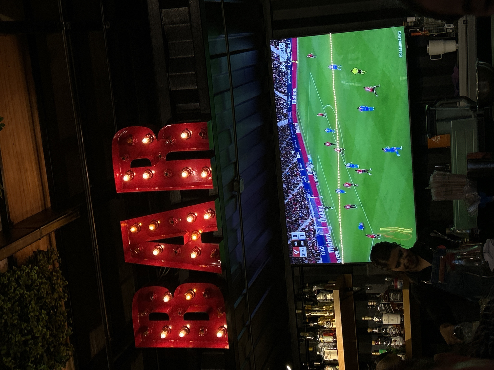 The scene is inside a bar, likely in Unknown, Unknown, indicated by the "BAR" sign illuminated in red. A television screen displays a live soccer game with the score OSASUNA 4, ATLETI 0 at 21:48. The game has branding and logos for "DIGI," "Mahou," "San Miguel," "LALIGA@uel," and "#OsasunaAtleti," along the sides. Below the TV, shelves are stocked with various bottles of liquor. A person stands behind the bar, possibly a bartender. The environment is dimly lit, with wood and metal elements visible in the bar's structure. A green bush is seen on the left side of the frame.