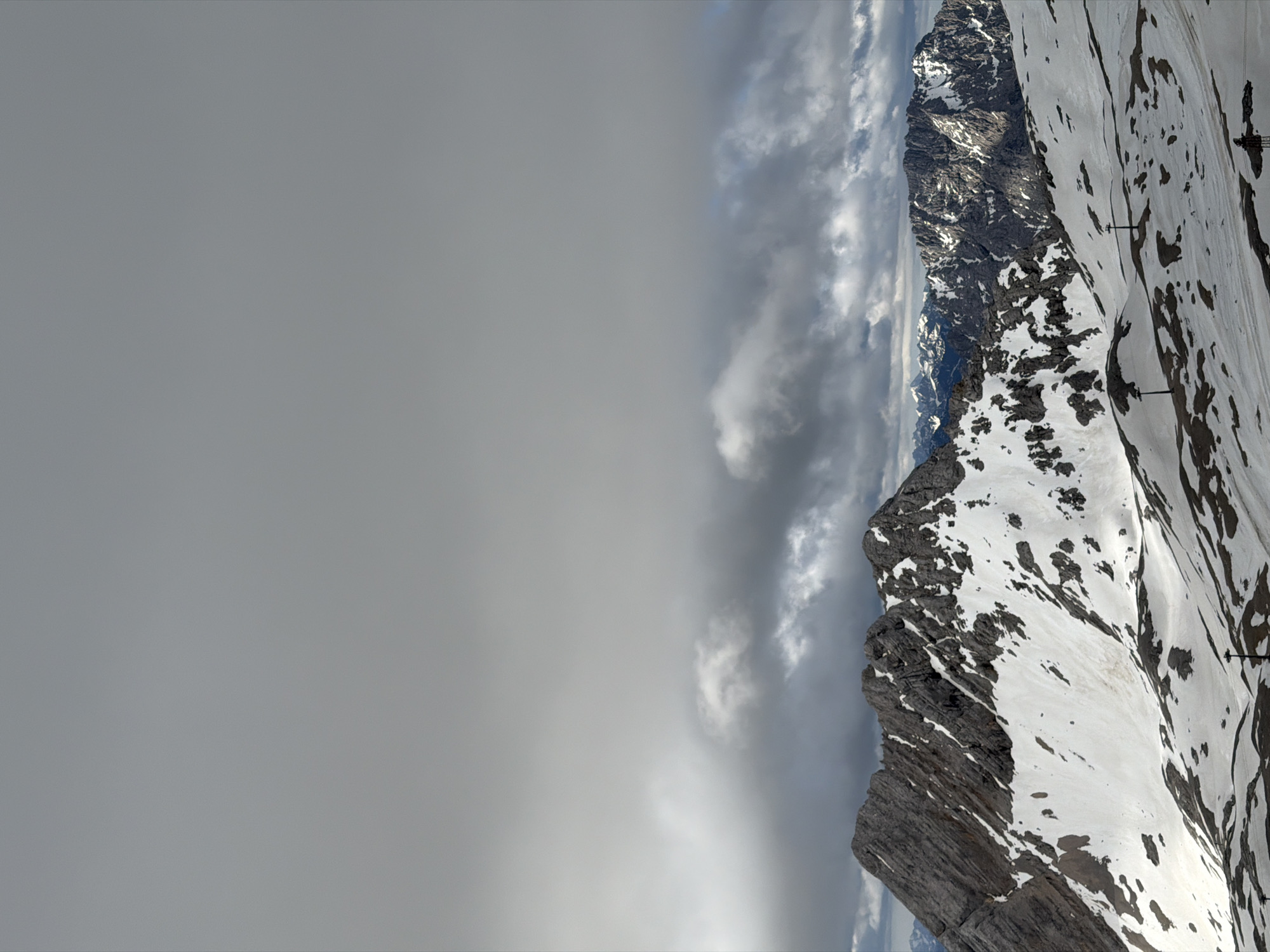 The image depicts a vast, overcast sky dominating the upper portion of the frame. Below the sky, a dramatic mountain range stretches across the landscape. The foreground is covered in snow, with patches of exposed rock and dark soil. Several ski lift towers are visible in the snowy terrain. The middle ground features more snow-covered mountains, some with jagged rocky peaks. In the distance, additional mountain ranges are visible, their forms softened by the atmospheric haze. The overall scene suggests a high-altitude, mountainous environment, likely the Bavarian Alps near Garmisch-Partenkirchen, Germany, given the presence of ski infrastructure and the characteristic alpine scenery. No people or specific actions are identifiable.