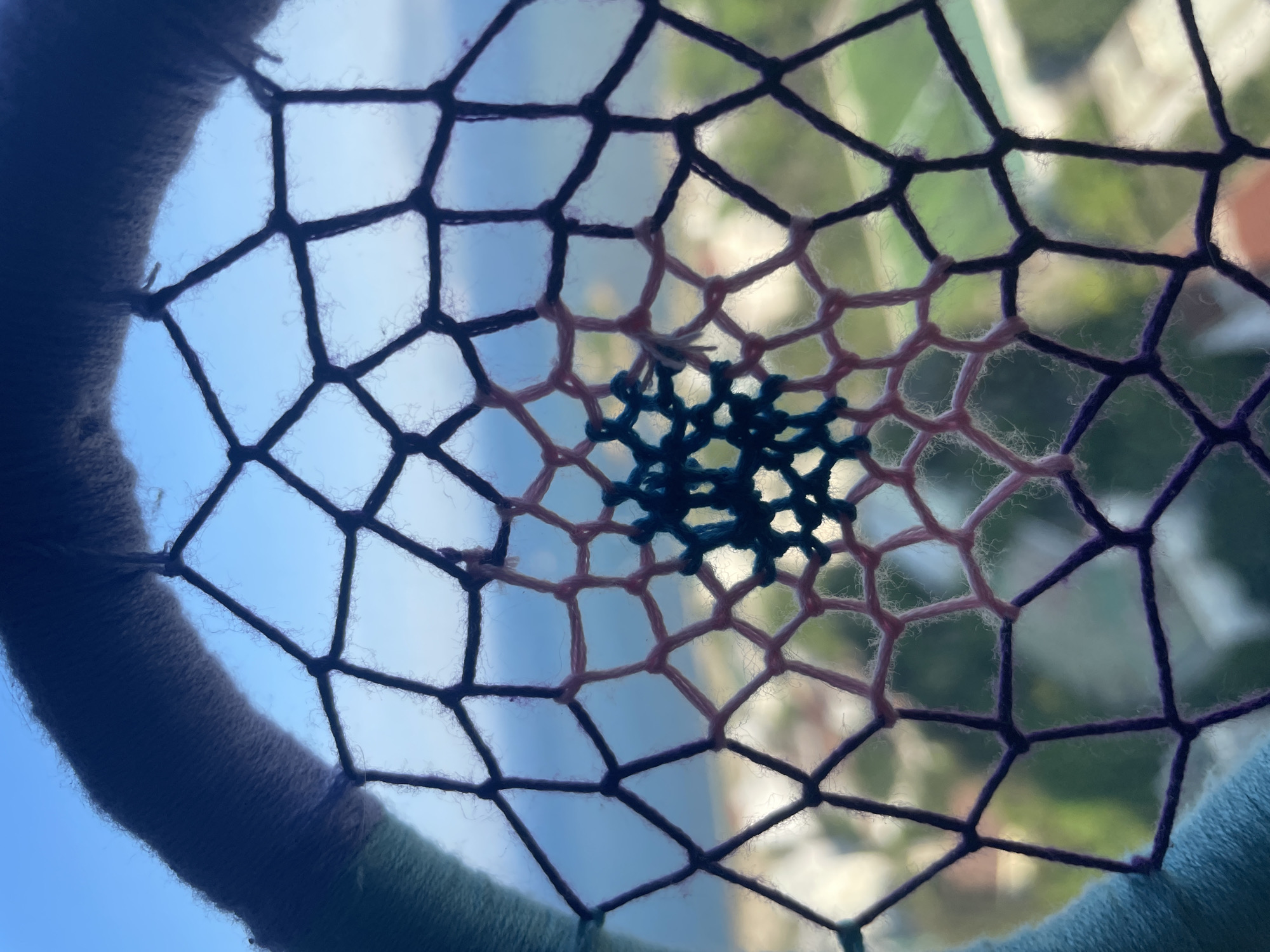 The image is a close-up of a dreamcatcher, viewed from behind, with a blurred background that appears to be an outdoor scene. The dreamcatcher has a circular hoop wrapped in fabric, transitioning from teal to a darker shade. A web of thread emanates from the hoop, with sections in black, light pink, and a central, more intricate pattern in dark teal. The background shows a hazy blue sky meeting a distant, blurred landscape with hints of green foliage and structures. The dreamcatcher is positioned in front of a window or opening, allowing the light and the outdoor environment to be visible through its open weave. There are no people or specific identifiable landmarks visible in the image.