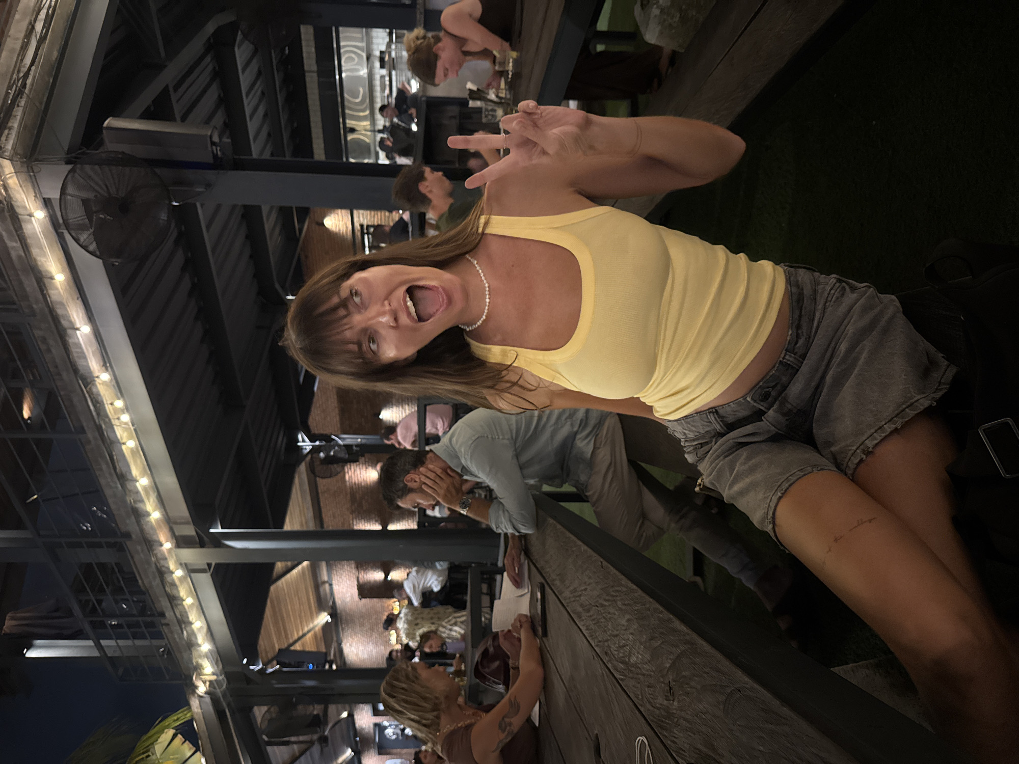 This sideways-oriented photograph captures a lively evening scene at an open-air restaurant or bar in North Kuta, Indonesia. The primary subject is a woman in the foreground with brown hair and bangs, wearing a bright yellow tank top, grey denim shorts, and a pearl necklace. She is depicted with a wide, open-mouthed expression of excitement or laughter, her hands raised playfully, one forming a "peace" sign.
The setting features an industrial-chic aesthetic, characterized by dark exposed metal beams supporting the roof structure. Strings of warm LED lights and ceiling fans are suspended from these beams, illuminating the space and indicating the nighttime setting. Wooden bar-height tables or railings are prominent in the foreground and midground, where several other patrons are visible. In the midground, a man in a light blue shirt leans against a railing, and a woman with blonde hair and a tattoo on her arm is seated at a table. These individuals appear to be socializing and relaxing, contributing to the convivial atmosphere.
The background shows more seating areas and a dark grassy patch, suggesting a spacious outdoor or semi-outdoor component to the venue. The overall ambiance is casual and social. A subtle, cursive tattoo is visible on the main subject's upper thigh, though the specific text is too small and indistinct to decipher clearly.