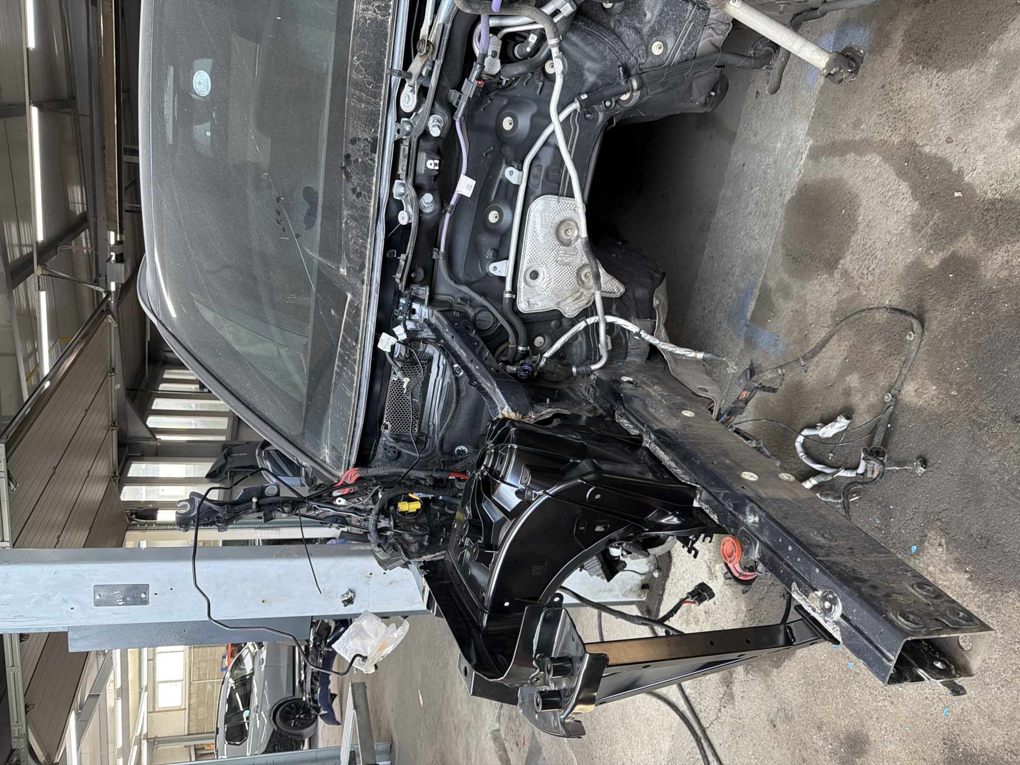 The image shows a partially disassembled car undergoing repairs, likely in a garage or repair shop. The front end of the vehicle is significantly dismantled, revealing the internal structure, wiring, and various components. The car is elevated on a lift, and other vehicles can be seen in the background, suggesting a professional automotive environment. The scene appears industrial, with a concrete floor and bright lighting. No people are visible in the frame. The specific location within the city of Unknown, Unknown is not discernible from the provided visual information.