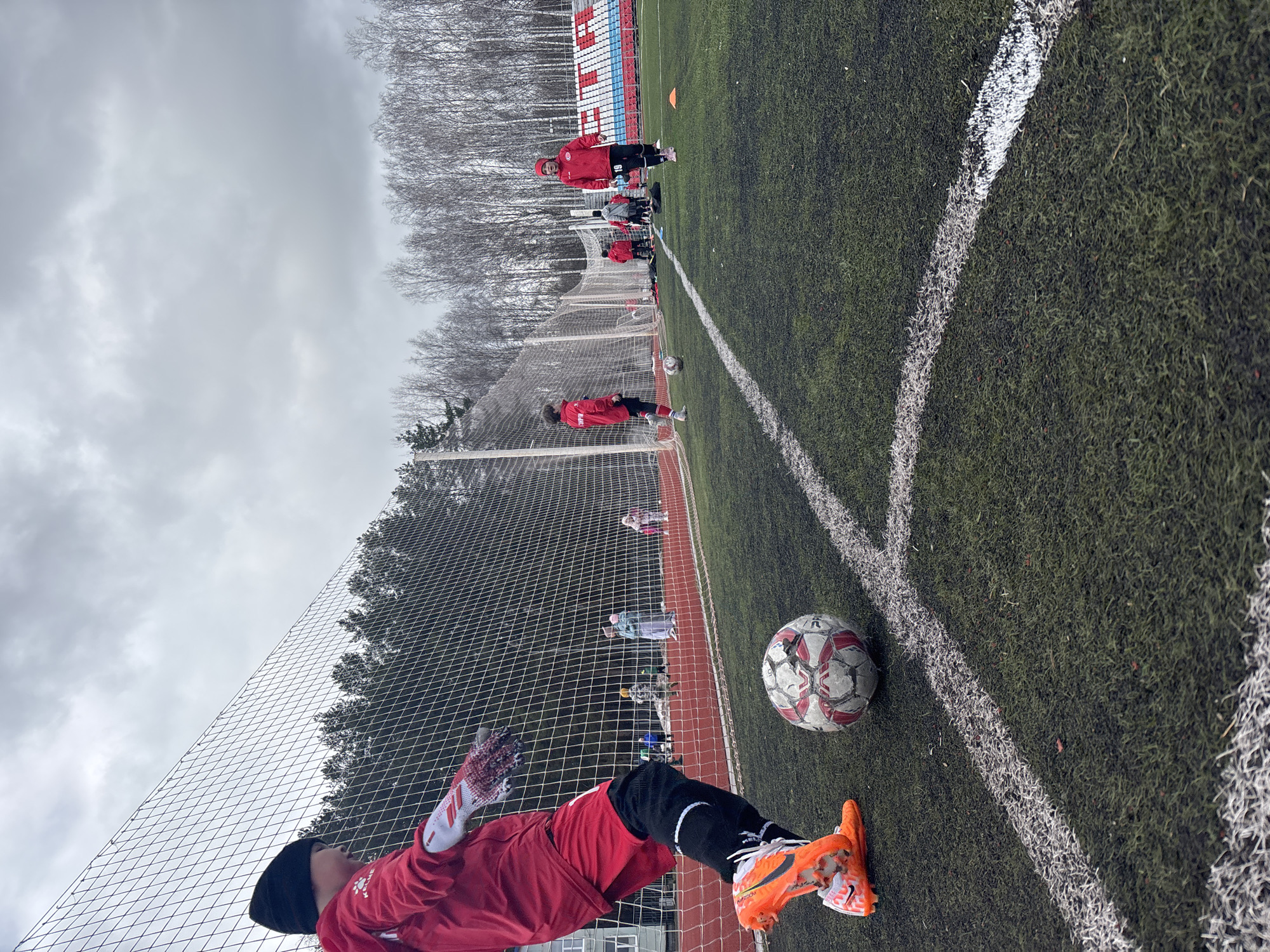 The image depicts a soccer field in Chaikovsky, Russia, on an overcast day. A soccer ball rests on the field near a corner marked by white lines. A soccer player, wearing a red jersey, black leggings, and orange cleats, is lying on the ground near the goal net. Other individuals in red jerseys are visible in the background, near the edge of the field, along with trees and a building with blue and red sections.