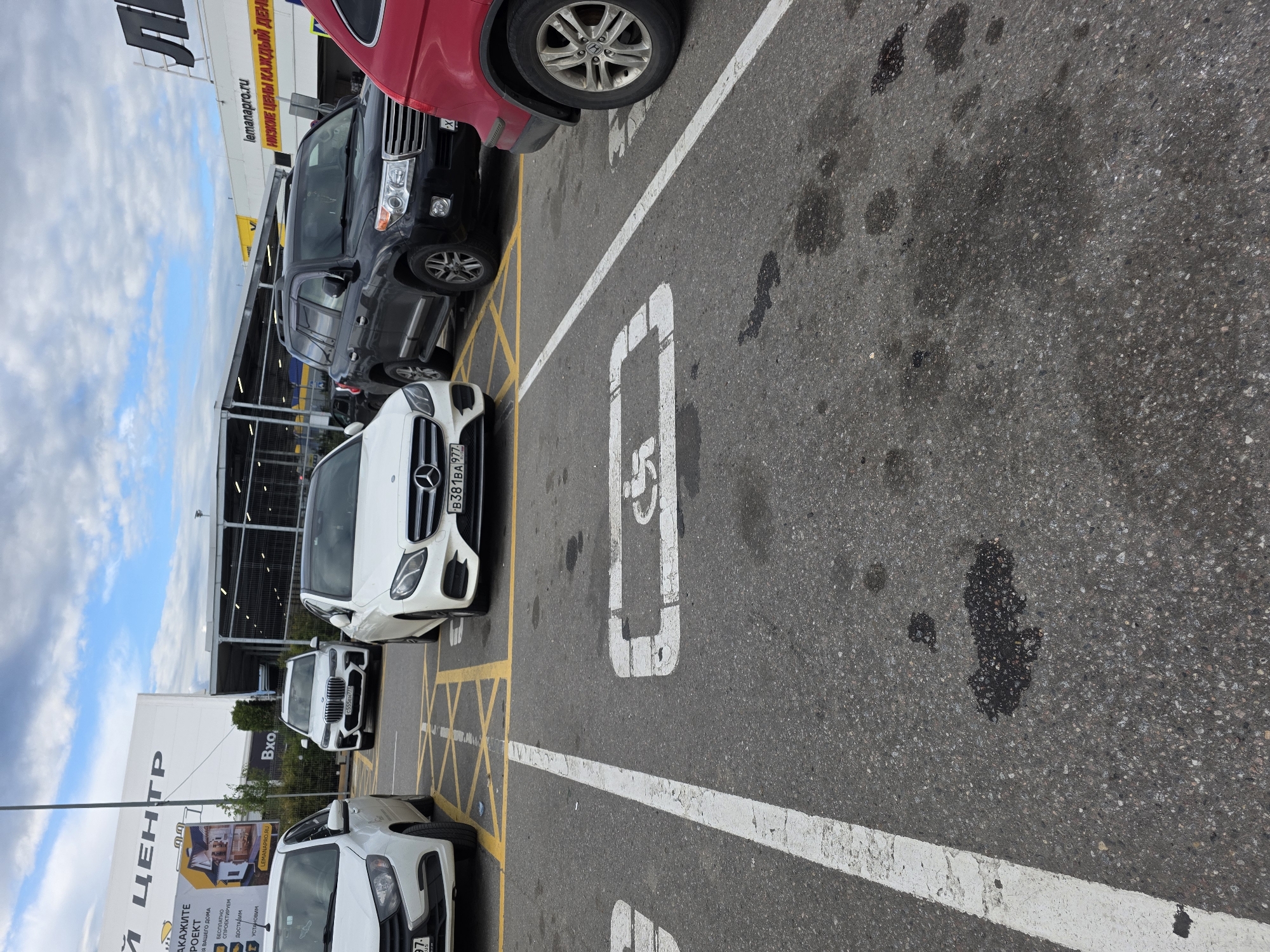 An exterior medium shot captures a parking lot scene. In the foreground, a white line divides the pavement. To the right of this line, a white square with a wheelchair symbol is painted on the asphalt. Further back, a white sedan is parked in a marked bay. Next to it is a white BMW SUV. To the right of the BMW is a black Toyota Land Cruiser. In the far right of the image, the rear of a red vehicle is partially visible. Above the vehicles, a large white building with a large blue and white sign is visible. The sky is partly cloudy.