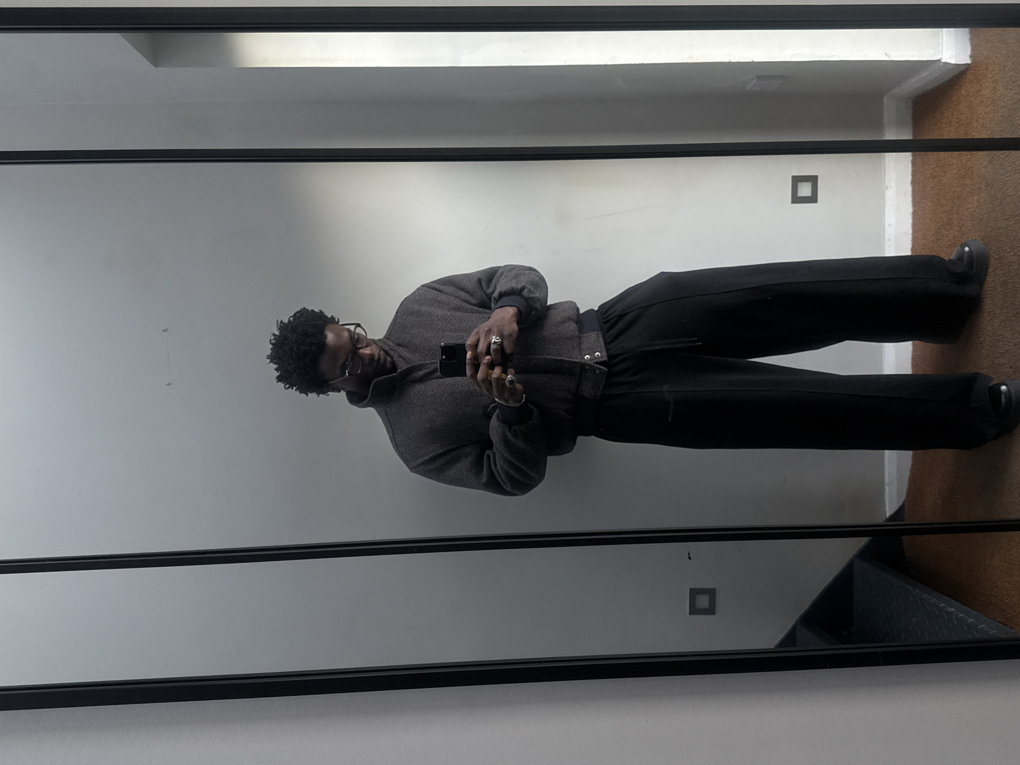 A person is reflected in a large, rectangular mirror with a black frame. The person has dark skin, dark curly hair, and is wearing glasses, a gray herringbone jacket, black pants, and black shoes. They are holding a smartphone and appear to be taking a selfie. The background shows a plain white wall with a square electrical outlet. The bottom right corner of the reflection reveals a glimpse of a brown carpet or floor. Based on the interior architecture, the location may be a modern residential or commercial space in London, United Kingdom.