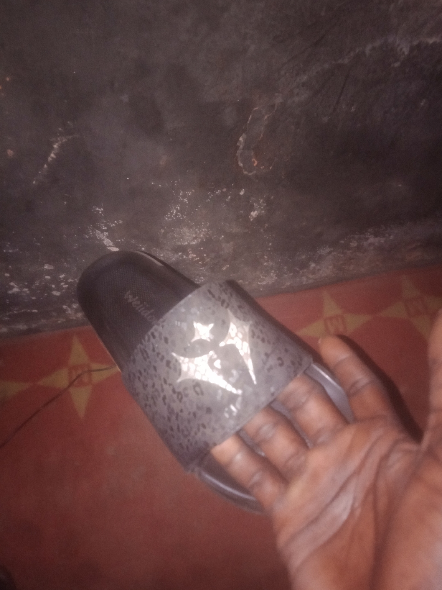 A person's hand is in the foreground, holding a slide sandal. The sandal is dark gray with a patterned strap featuring abstract shapes that appear white. The sole of the sandal is visible, and it has a textured pattern. The background shows a dark, textured surface above and a reddish-orange tiled floor below. The lighting is dim, casting shadows. The scene appears to be indoors, but the exact location is not discernible.