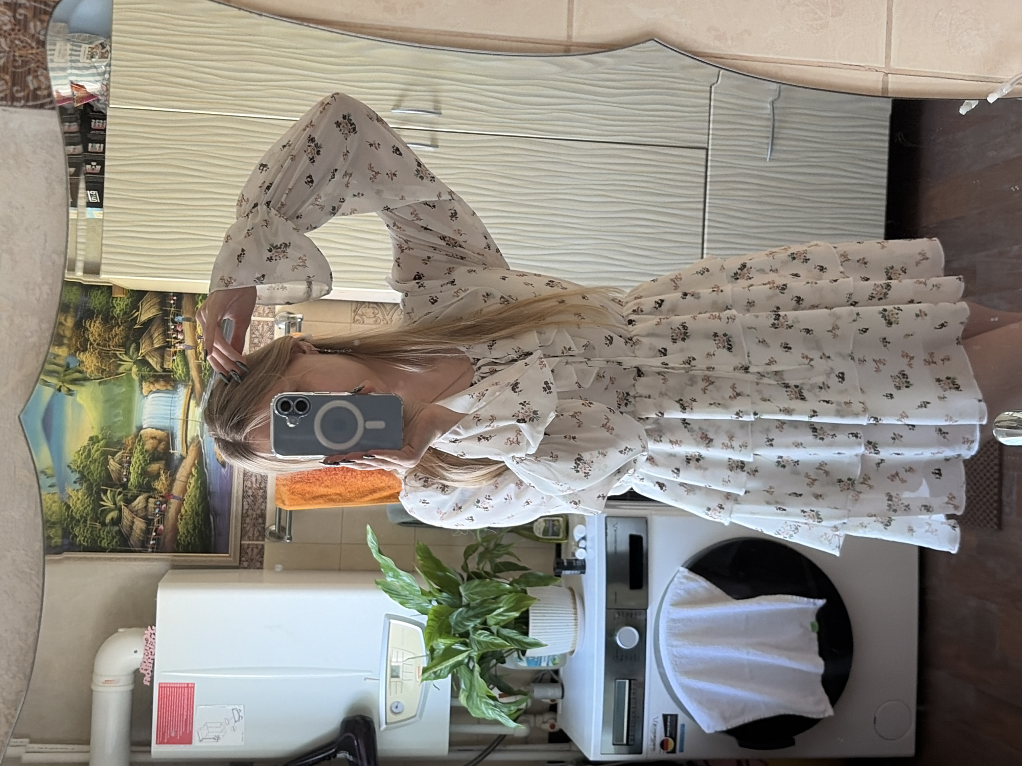 A woman is visible in a mirror reflection, holding a phone up to take a picture of herself. She is wearing a white, floral-patterned, ruffled dress with long sleeves. The environment includes a bathroom setting. Behind her in the reflection is a painting, a white water heater, and a washing machine with a white cloth on top. A potted plant is also visible between the water heater and the washing machine. Above, cabinets are visible. The city is Unknown, Unknown.