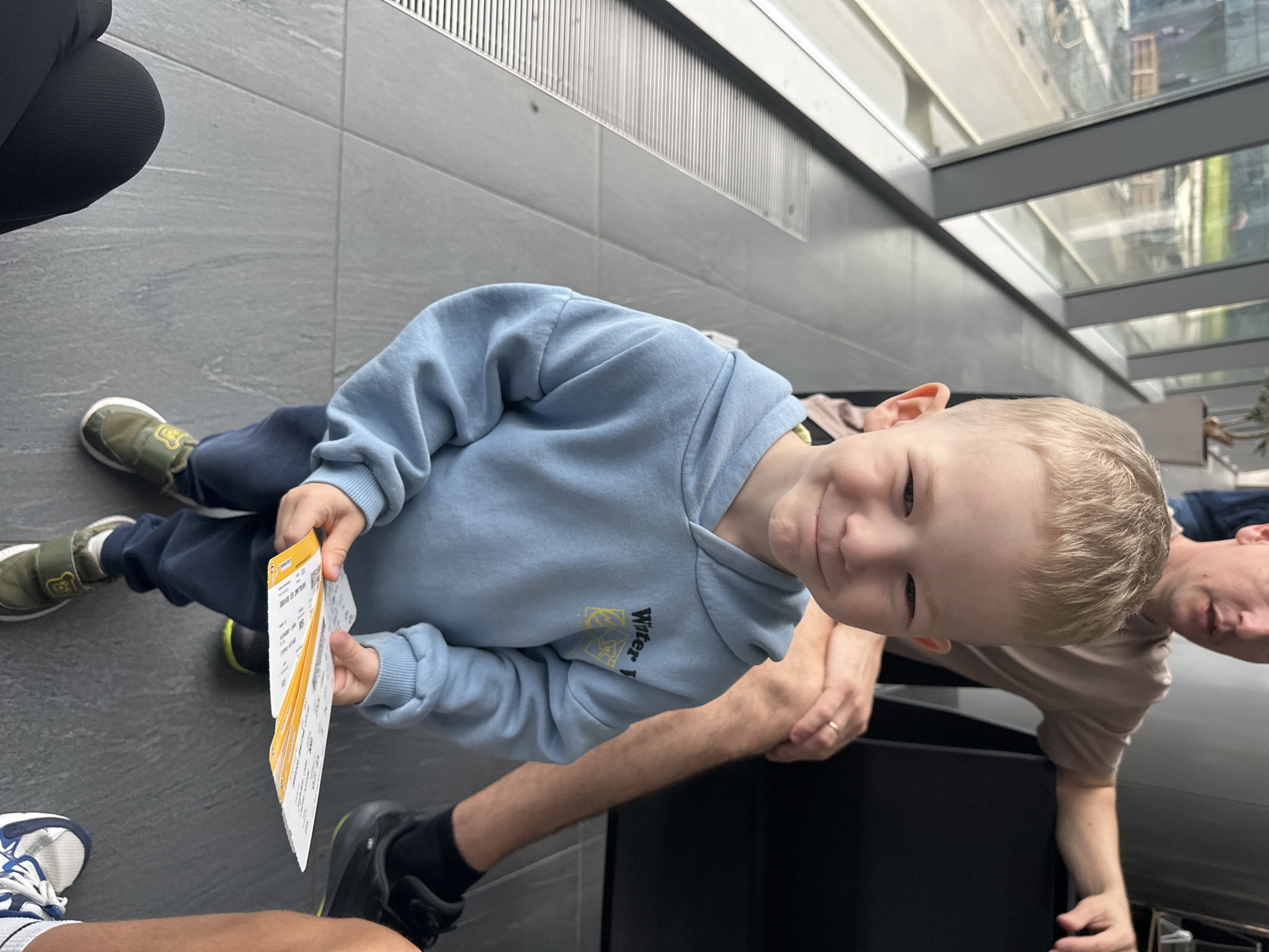 A young boy with blond hair and blue eyes stands in what appears to be an airport waiting area, holding several boarding passes. He is wearing a light blue hooded sweatshirt and dark blue pants. His stance suggests he might be waiting for a flight, given the boarding passes in his hand. In the background, a man, presumably his father, is seated, with his arm resting on the boy's shoulder. The environment features large windows, likely overlooking a tarmac or runway, and tiled flooring, common in airport terminals. The specific location is identified as the Bolshoye Savino airport in Russia, suggesting the boy and his family are either arriving or departing from this city.