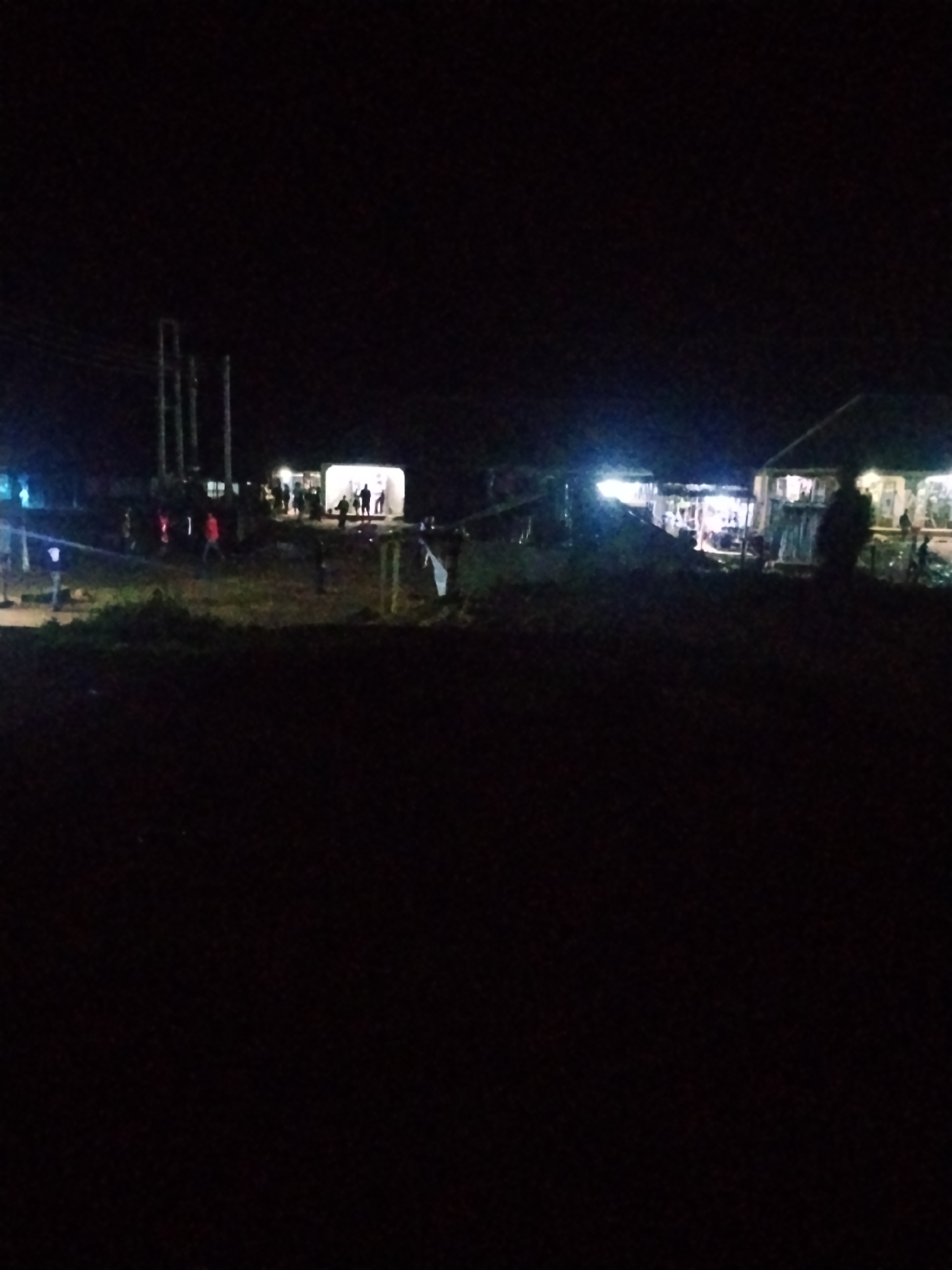 This image depicts an outdoor scene at night in Durimi, Nigeria. The foreground is dark, with the middle ground showing several illuminated structures, possibly shops or stalls, with people gathered around them. There are also utility poles and wires visible in the background, indicating the presence of infrastructure. The lighting is artificial and appears to be from overhead bulbs or lamps. The overall impression is of a moderately populated area at night, likely a marketplace or gathering spot. The lighting is dim and the image quality is poor, making precise identification of individuals or objects difficult.