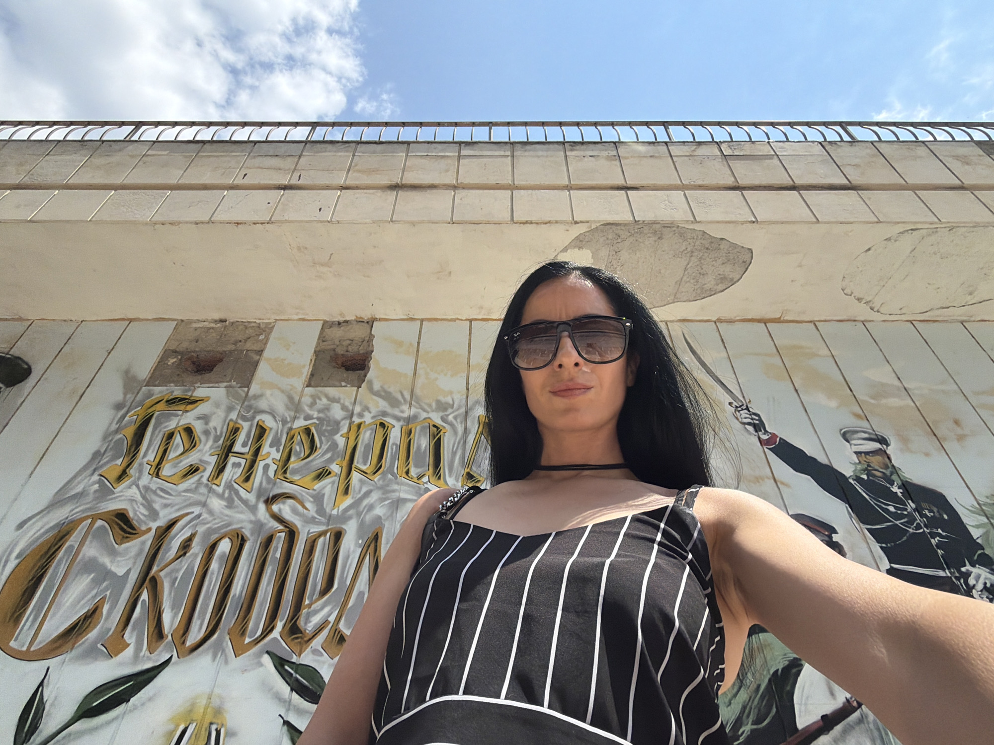 A low-angle, wide shot captures a woman standing in front of a mural. She is wearing a black and white striped tank top, large sunglasses, and a thin black choker necklace. Her long, dark hair is down. The mural behind her depicts historical figures in military attire, with one figure on the right holding a sword aloft. Golden Cyrillic text is visible on the left side of the mural. The scene appears to be outdoors on a sunny day, with a blue sky and scattered clouds overhead. The wall the mural is on is made of concrete blocks, with some areas showing wear and tear. The overall impression is a casual outdoor portrait against a backdrop of historical art. The time of day appears to be daytime, likely late morning or early afternoon, given the bright sunlight casting shadows. The location is suggested to be Pleven, Bulgaria, based on the presence of Cyrillic text, likely referencing historical figures or events relevant to the region.