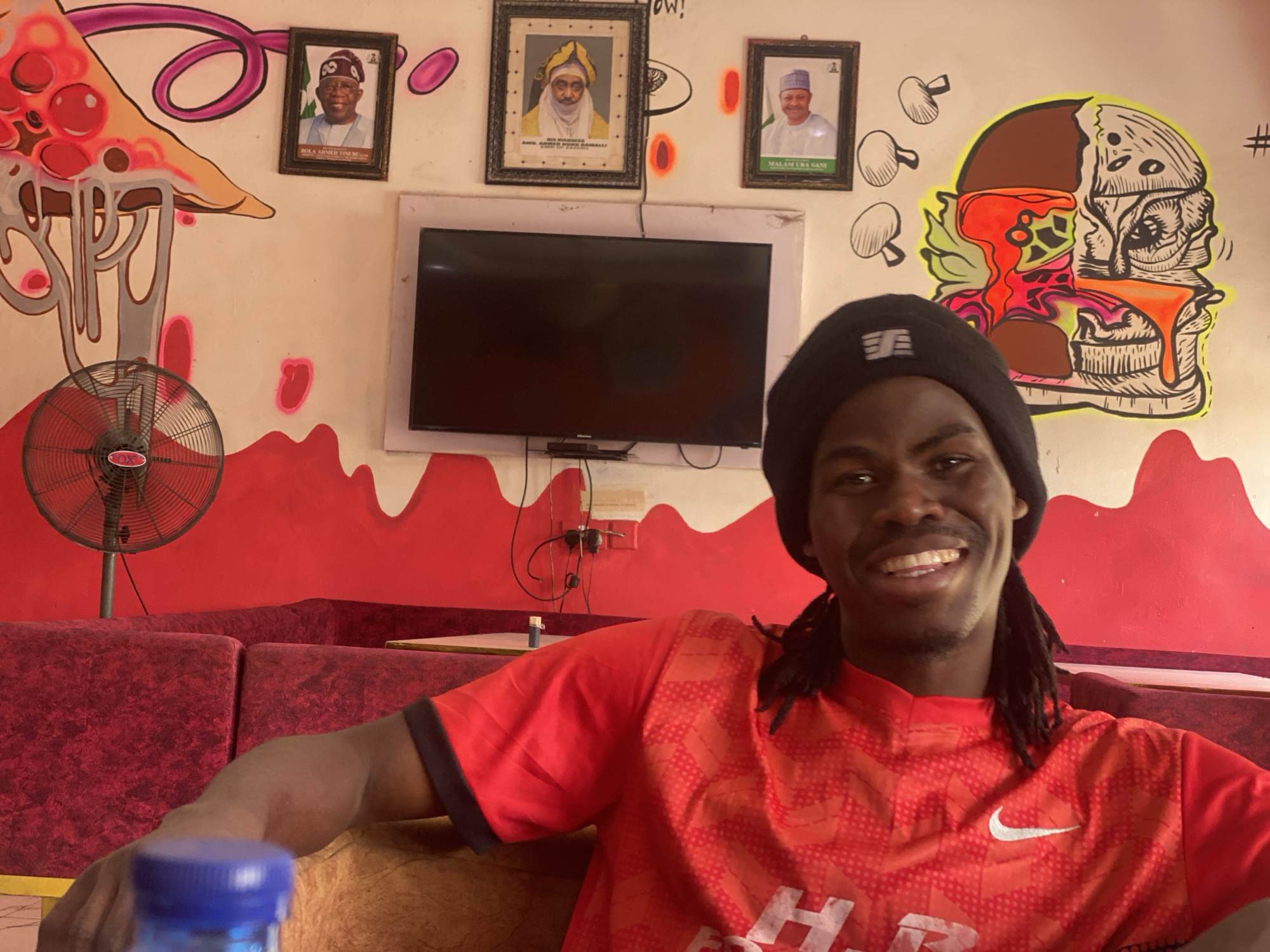 A smiling man wearing a black beanie and a red patterned Nike t-shirt sits in a restaurant or lounge. He has dark skin and dreadlocks. A bottle with a blue cap sits in the foreground.
Behind him, the wall is painted with red drips and cartoonish drawings of a pizza slice and a hamburger. There are also three framed portraits of men in traditional dress. A flat-screen television hangs on the wall between the portraits and the cartoons. A standing fan is on the left, and a red sofa is visible.
The restaurant or lounge is located in Tukur Tukur, Nigeria.
