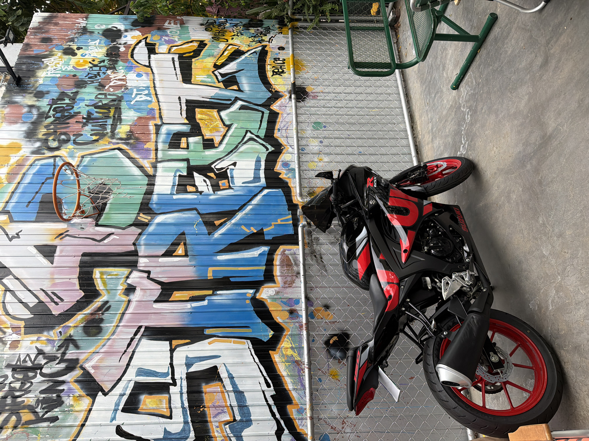 A black and red Suzuki GSX motorcycle is parked on a concrete surface in front of a chain-link fence. Behind the fence, a corrugated metal wall is covered in vibrant graffiti. A basketball hoop with a net is mounted on the wall. To the right of the motorcycle, a green metal bench is situated on the concrete. The scene is outdoors, with a hint of foliage visible to the right. This appears to be an urban setting.