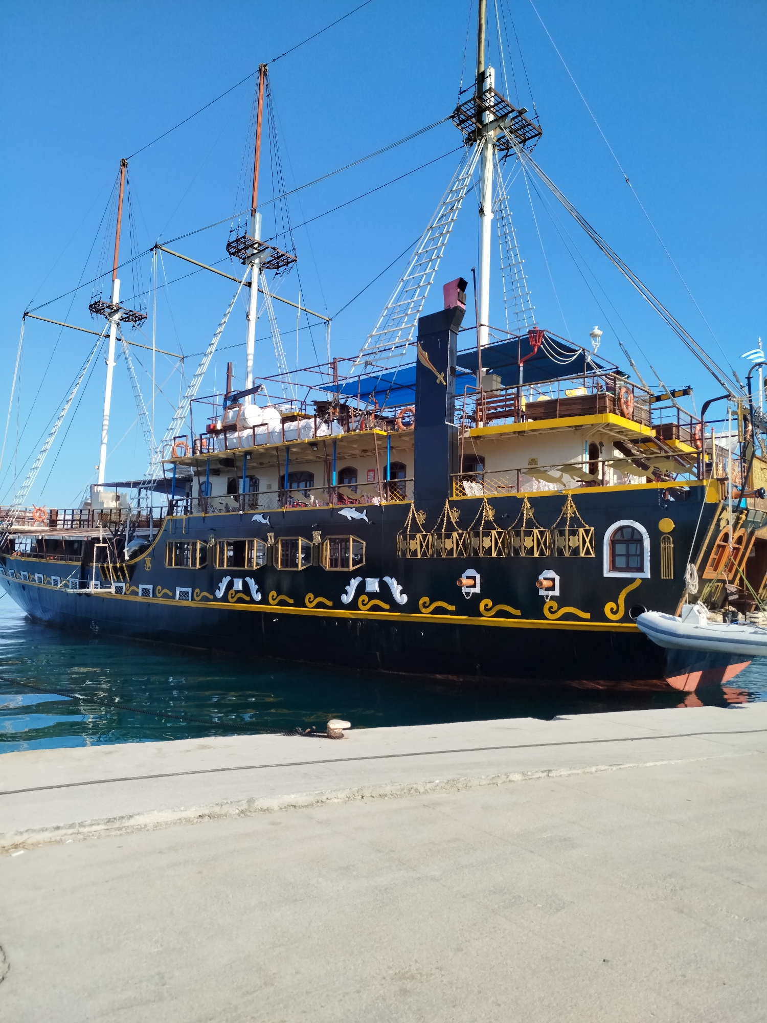 A black, multi-decked pirate-themed ship is docked at a concrete pier under a clear blue sky. The ship features multiple masts with rigging, and decorative yellow and white embellishments along its hull, including images of dolphins and other sea creatures. Several windows with ornate frames are visible. A small, inflatable boat is tied to the side of the larger ship. The water surrounding the ship is a deep blue-green, and the concrete pier is visible in the foreground. A mooring bollard is present on the pier. The scene suggests a daytime setting in a harbor or marina. The image appears to be taken in the Sithonia Municipal Unit, Greece, known for its coastal beauty and maritime activities.