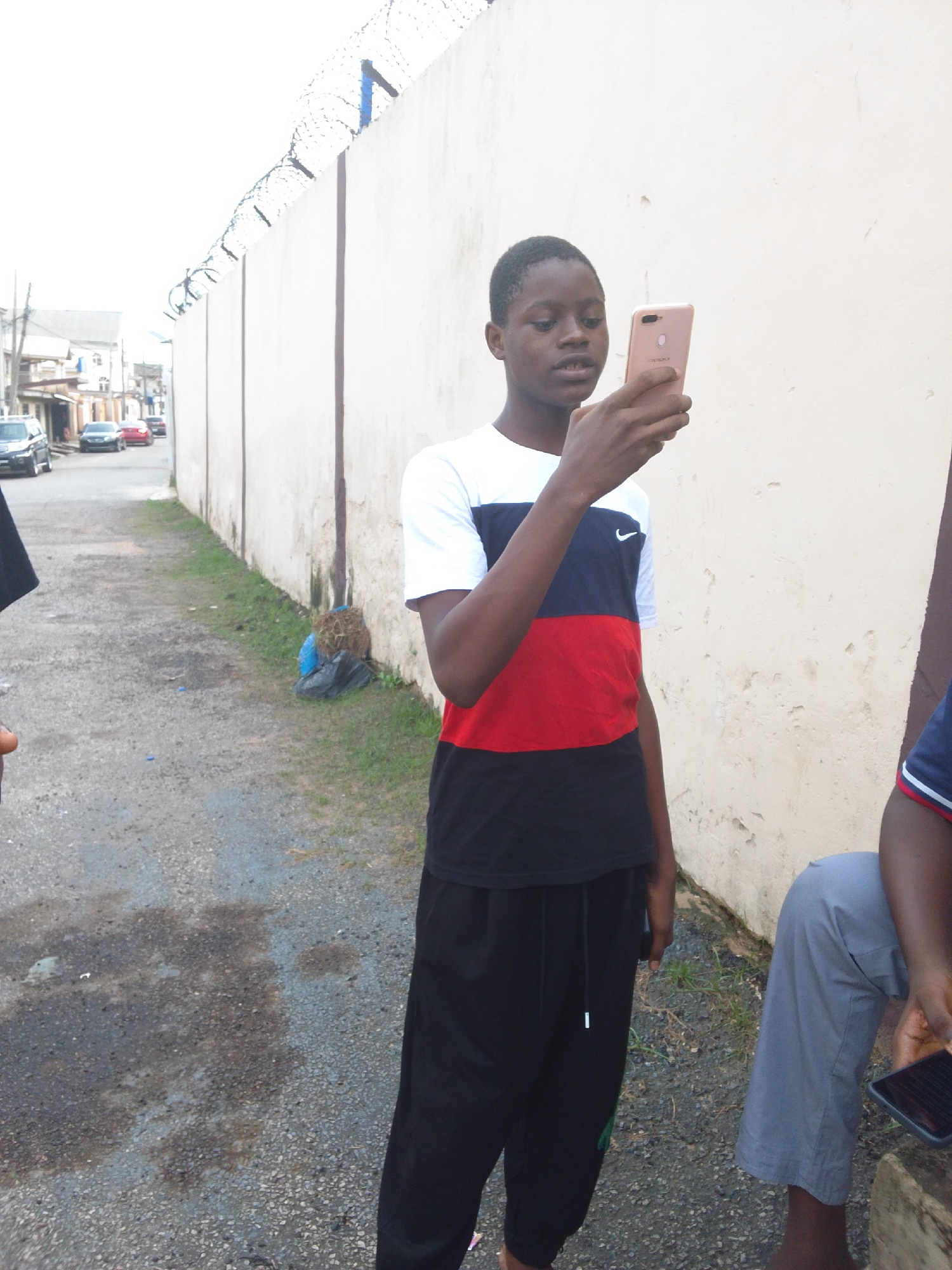 The image shows a young person standing in an outdoor setting, likely a street or alley in an urban area, potentially Shangisha, Nigeria. The individual is holding a mobile phone in their hands and appears to be looking at the screen. They are wearing a short-sleeved t-shirt with a color-blocked design of white, blue, red, and black, along with dark-colored pants. To the right of this individual, another person is partially visible, wearing a blue shirt and gray shorts, also looking at a phone.
The background features a tall, light-colored wall topped with barbed wire. The wall lines the side of the street or alley. The ground is paved and shows some signs of discoloration or dampness. There are also cars visible in the distance on the left side of the frame. Debris, possibly trash, is visible near the wall.