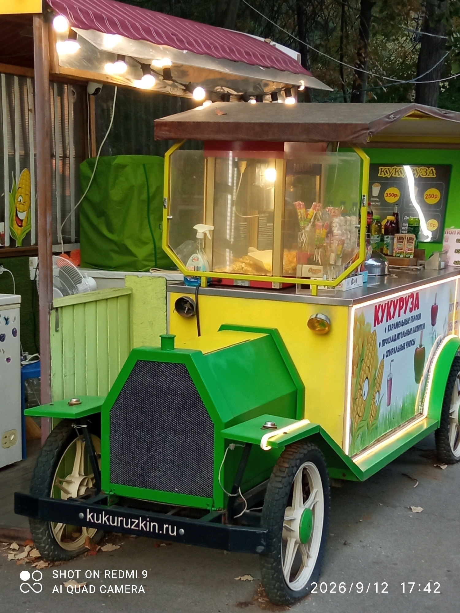A bright green and yellow food cart, designed to resemble an old-fashioned automobile, is parked on an asphalt surface. The cart is decorated with images of corn and features a popcorn machine. The front of the cart has a black grill and large wheels. The words "kukuruzkin.ru" are visible on a black bumper. The background includes trees, a corrugated metal fence, and a green tarp. A security camera is mounted on the overhang of the cart. The image is time-stamped with "2026/9/12 17:42" and includes a "SHOT ON REDMI 9 AI QUAD CAMERA" watermark. The text on the cart is in Russian and appears to advertise corn-based products and drinks. The scene is outdoors, likely in a public space.