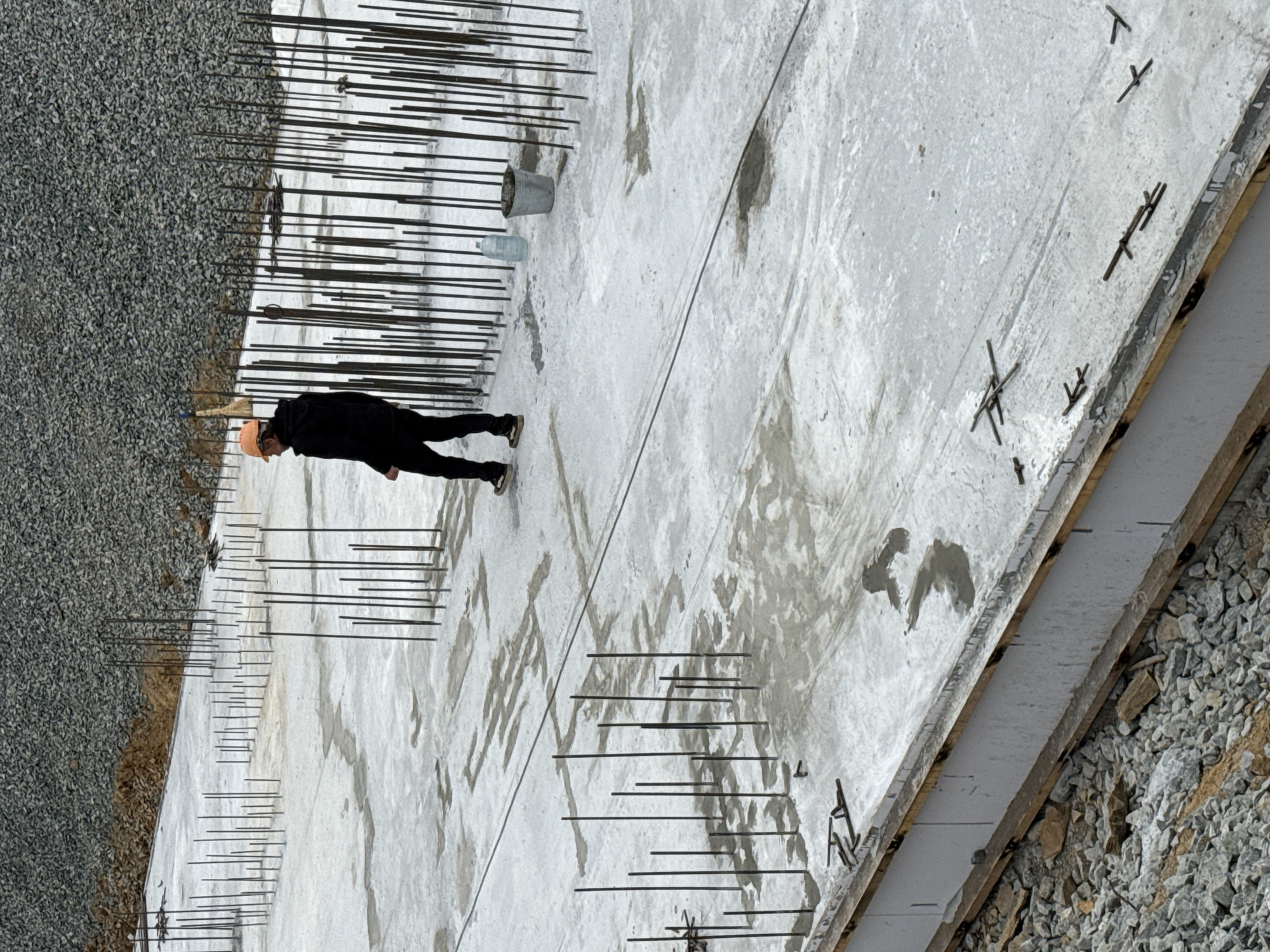The image shows a construction site, possibly in Leninsk, Russia. A person wearing a hard hat and dark clothing stands on a concrete slab with reinforcing steel bars protruding vertically. The slab is bordered by gravel and has a styrofoam edge. A metal bucket and a water bottle are also visible on the slab. The environment suggests an ongoing or recently completed phase of concrete construction.