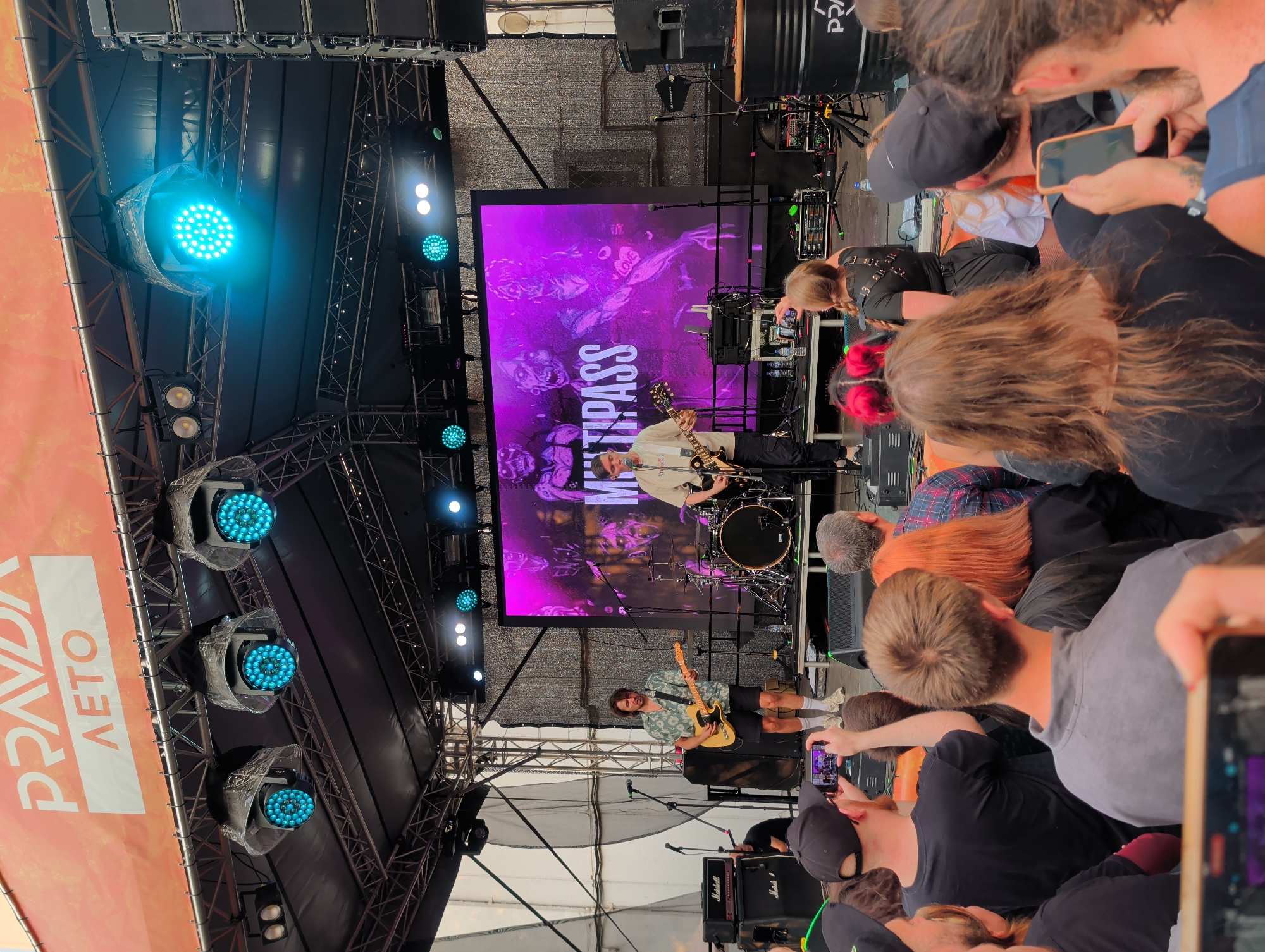 This image depicts an outdoor music concert stage. In the background, a large LED screen displays a purple graphic with skeletal figures and the word "MULTIPASS." Two male musicians are performing on stage; one is playing a yellow electric guitar and wearing a light green short-sleeved patterned shirt and black shorts, while the other is playing a dark electric guitar and wearing a light-colored t-shirt and black pants. A drum kit is visible behind them. Overhead, a stage rig holds multiple lighting fixtures, some emitting bright blue light. The stage is set up outdoors, and the audience is visible in the foreground, with many people holding up their phones to record or photograph the performance. A banner with the Cyrillic text "ПРАВДА ЛЕТО" (Pravda Leto - Truth Summer) is hung above the stage. The overall atmosphere suggests a lively music festival or concert event.