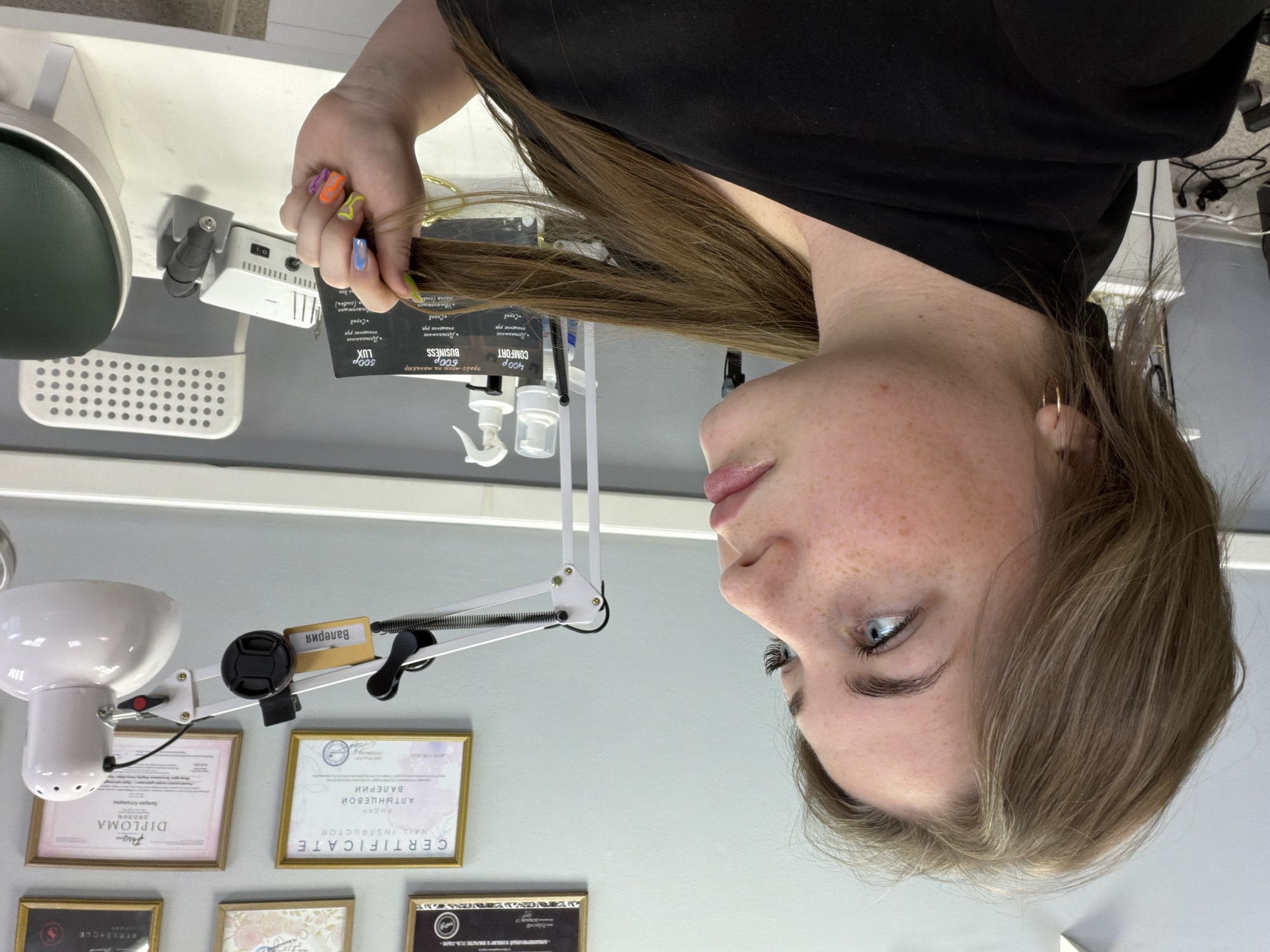 A young woman with shoulder-length brown hair and blue eyes is captured from the shoulders up, looking to her right. She has freckles across her nose and cheeks and is wearing a black top and small hoop earrings. Her left hand, adorned with colorful, abstract nail art, is holding a section of her hair. In the background, a white lamp with an adjustable arm is mounted on a wall. The wall itself is light gray and features several framed certificates and diplomas, including one labeled "CERTIFICATE NAIL INSTRUCTOR." To the right, a white chair and a white nail drill machine are visible on a white table. A menu with prices for manicure services is also displayed on the table. The overall setting appears to be a nail salon or a similar beauty establishment.